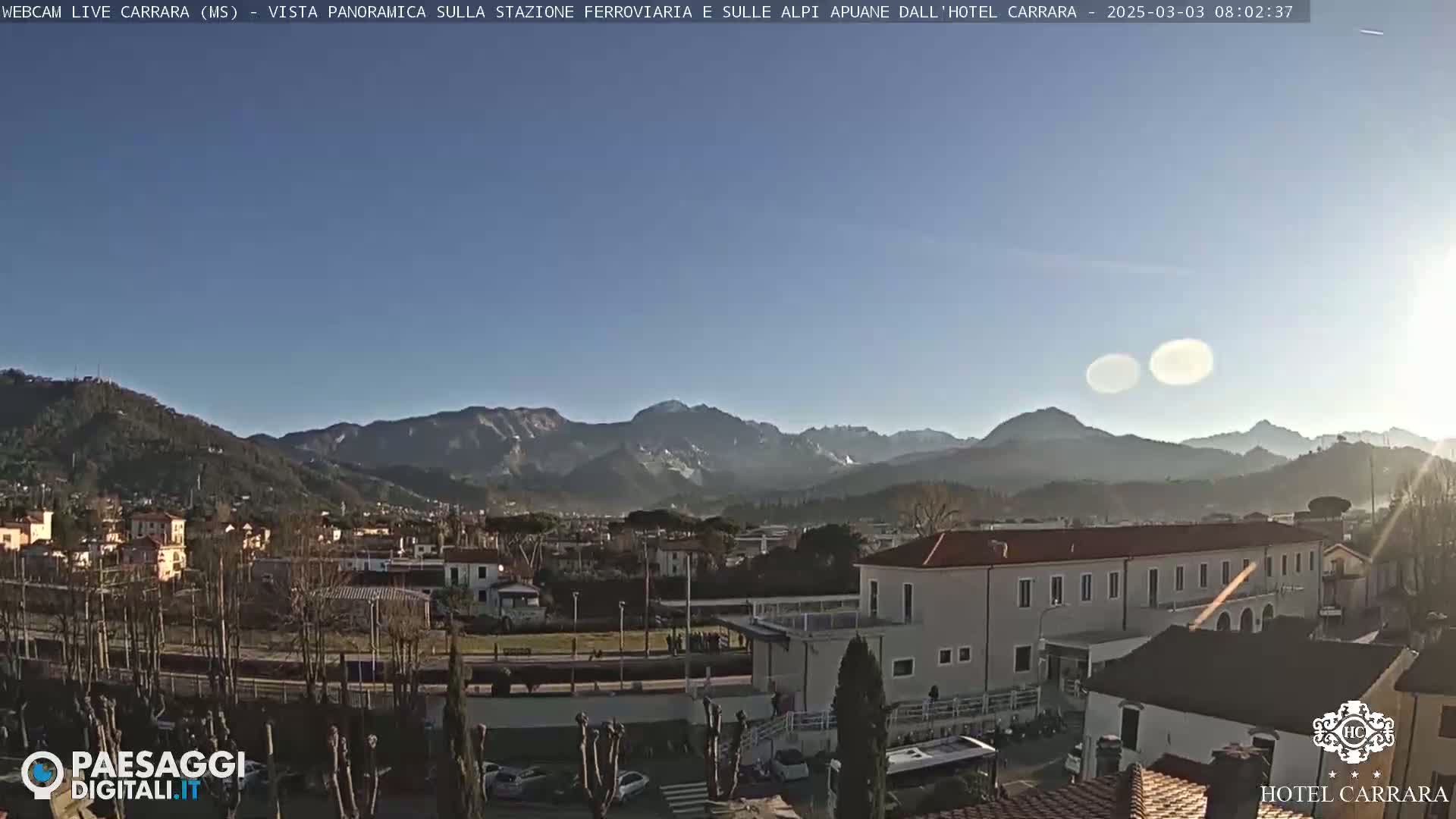 A panoramic view of a town nestled in a valley, with snow-capped mountains in the background under a clear, sunny sky.