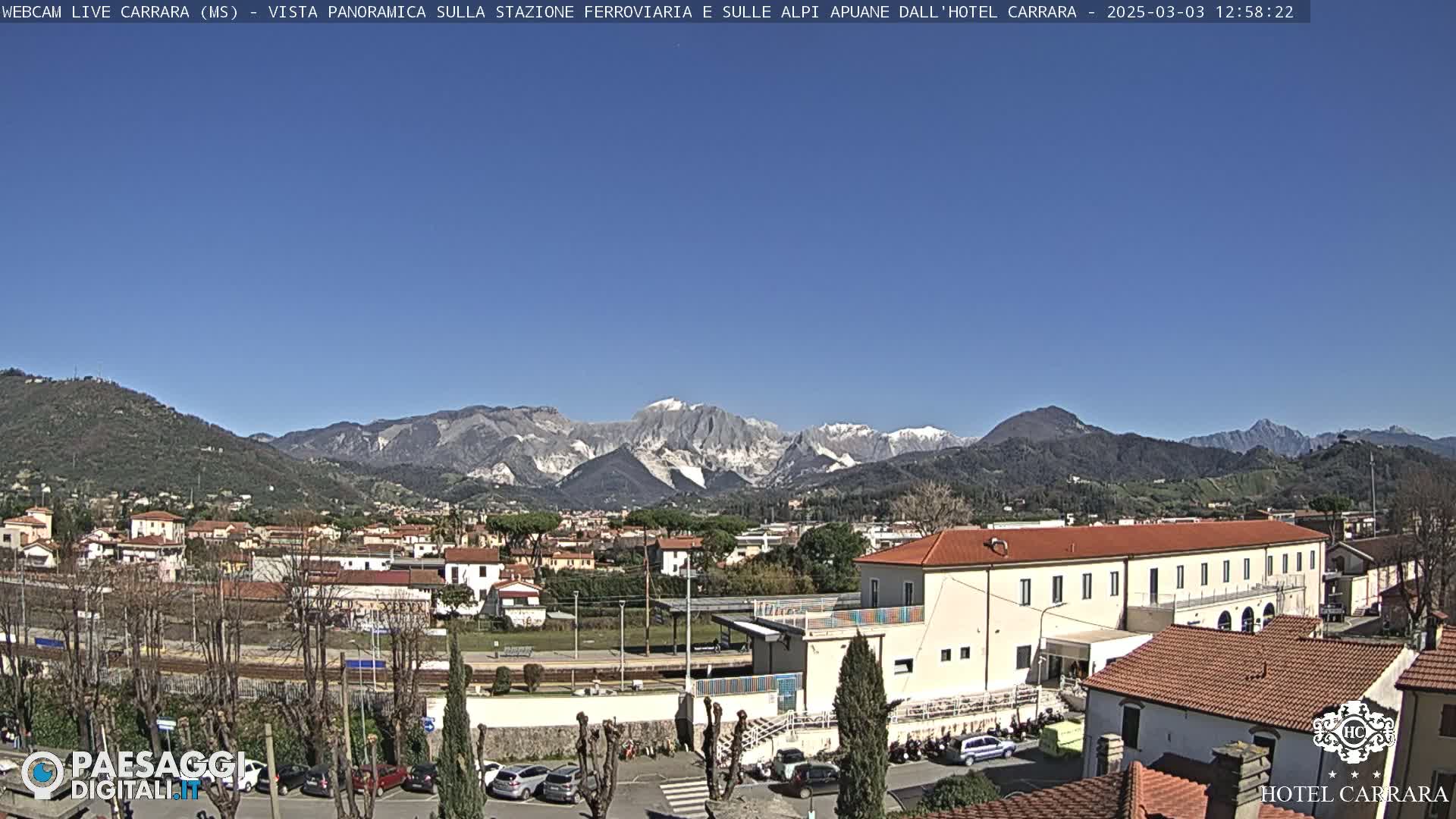 A clear, sunny day reveals a cityscape nestled in a valley, with snow-capped mountains in the background.