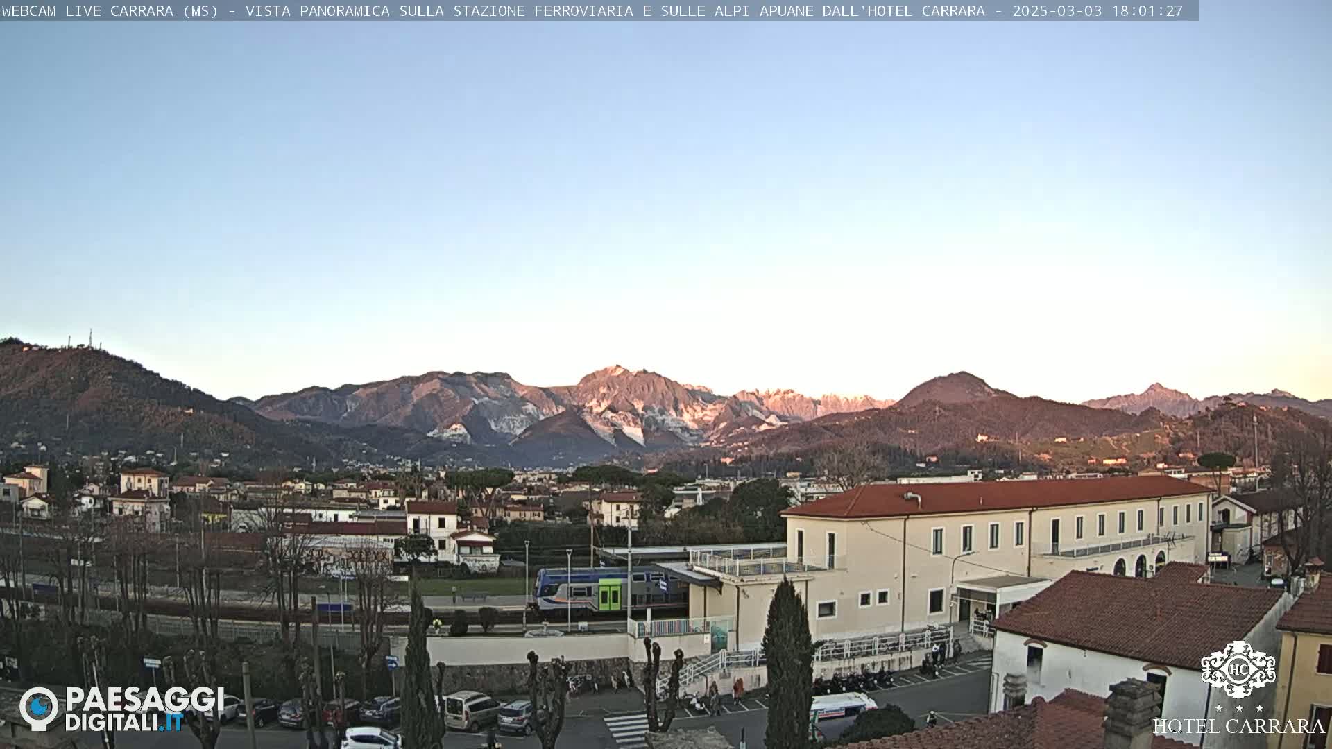 A long, low building sits near a train in a town nestled at the foot of snow-capped mountains under a clear, pale blue sky.