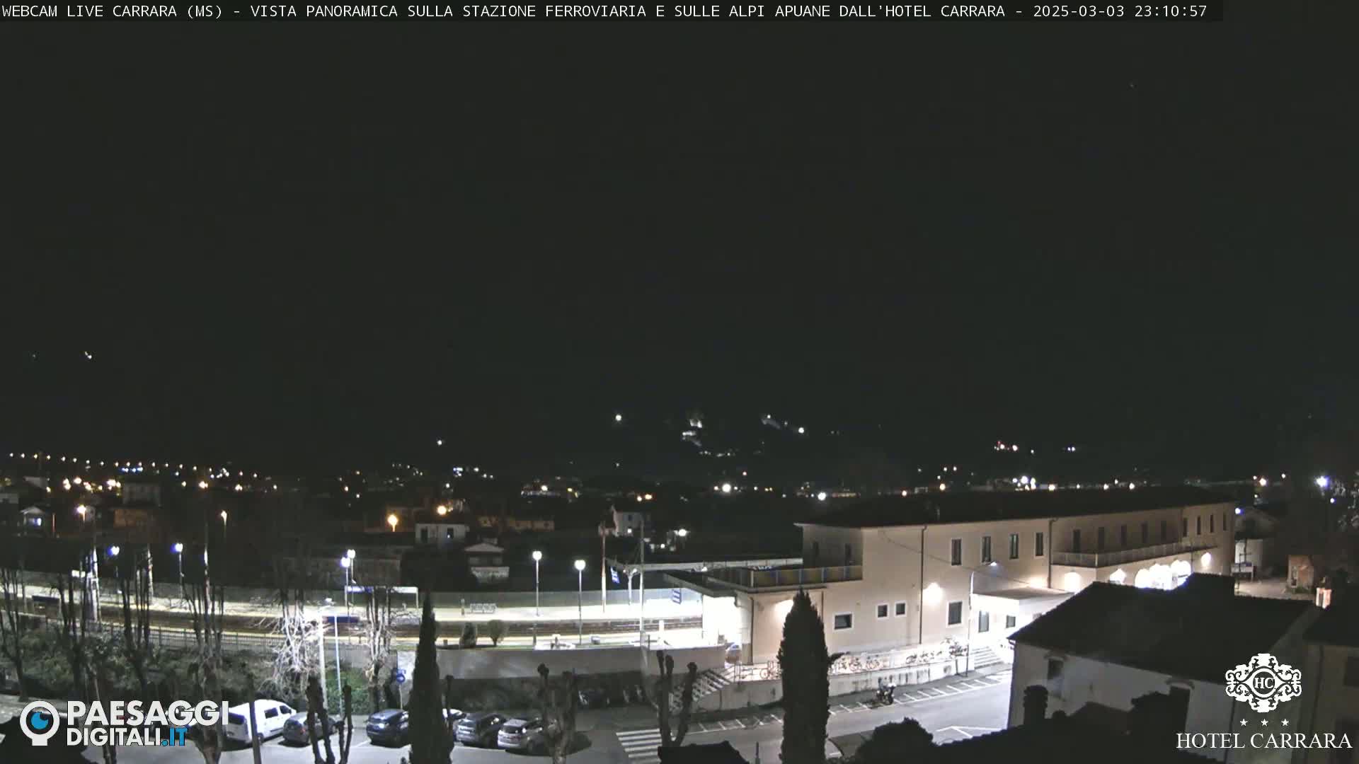 A nighttime, panoramic view of a town's outskirts, featuring a train station and a large building, under a clear, dark sky.