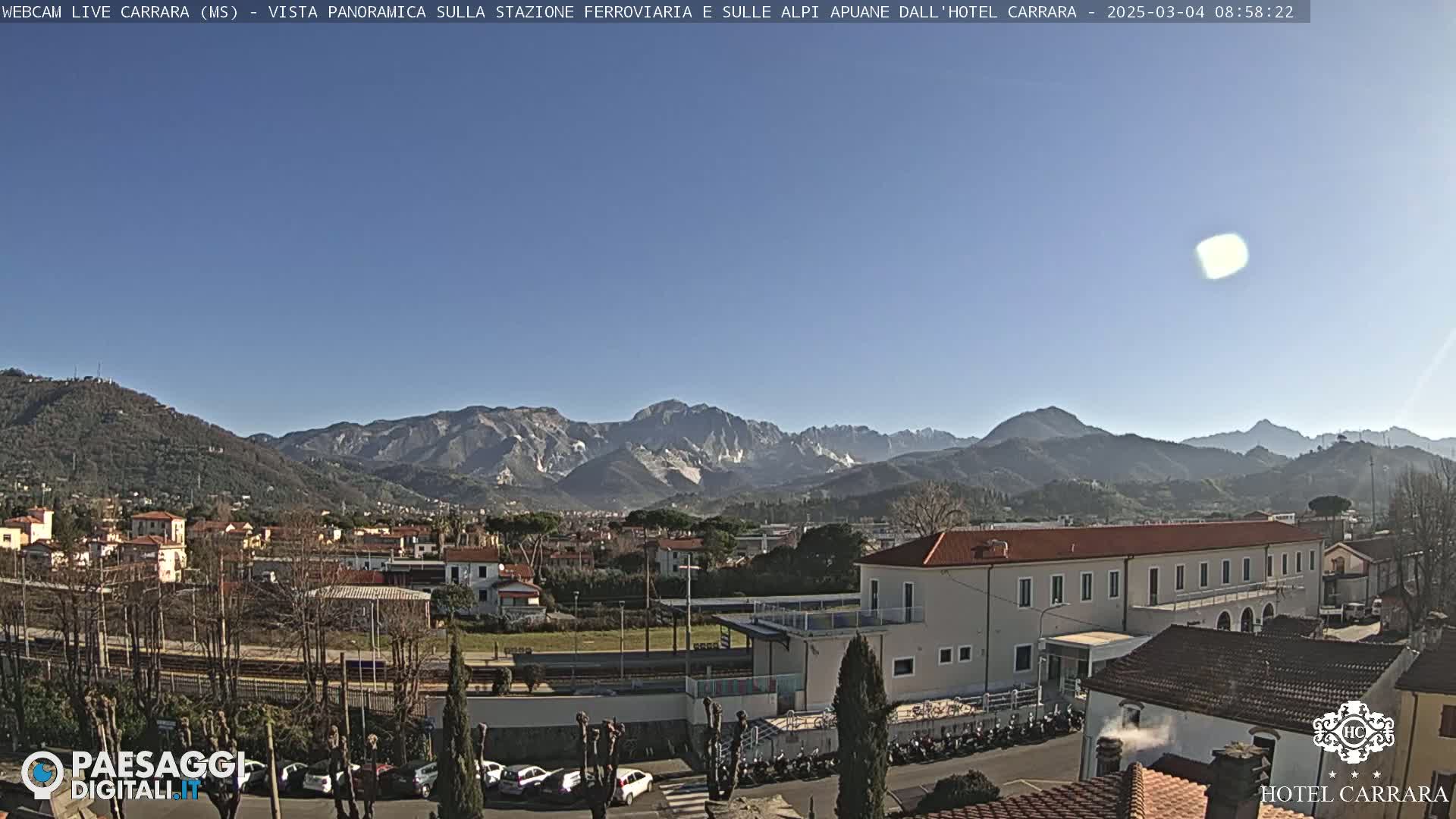 A long, low building sits in a town nestled in a valley, with a mountain range visible in the background under a clear, sunny sky.