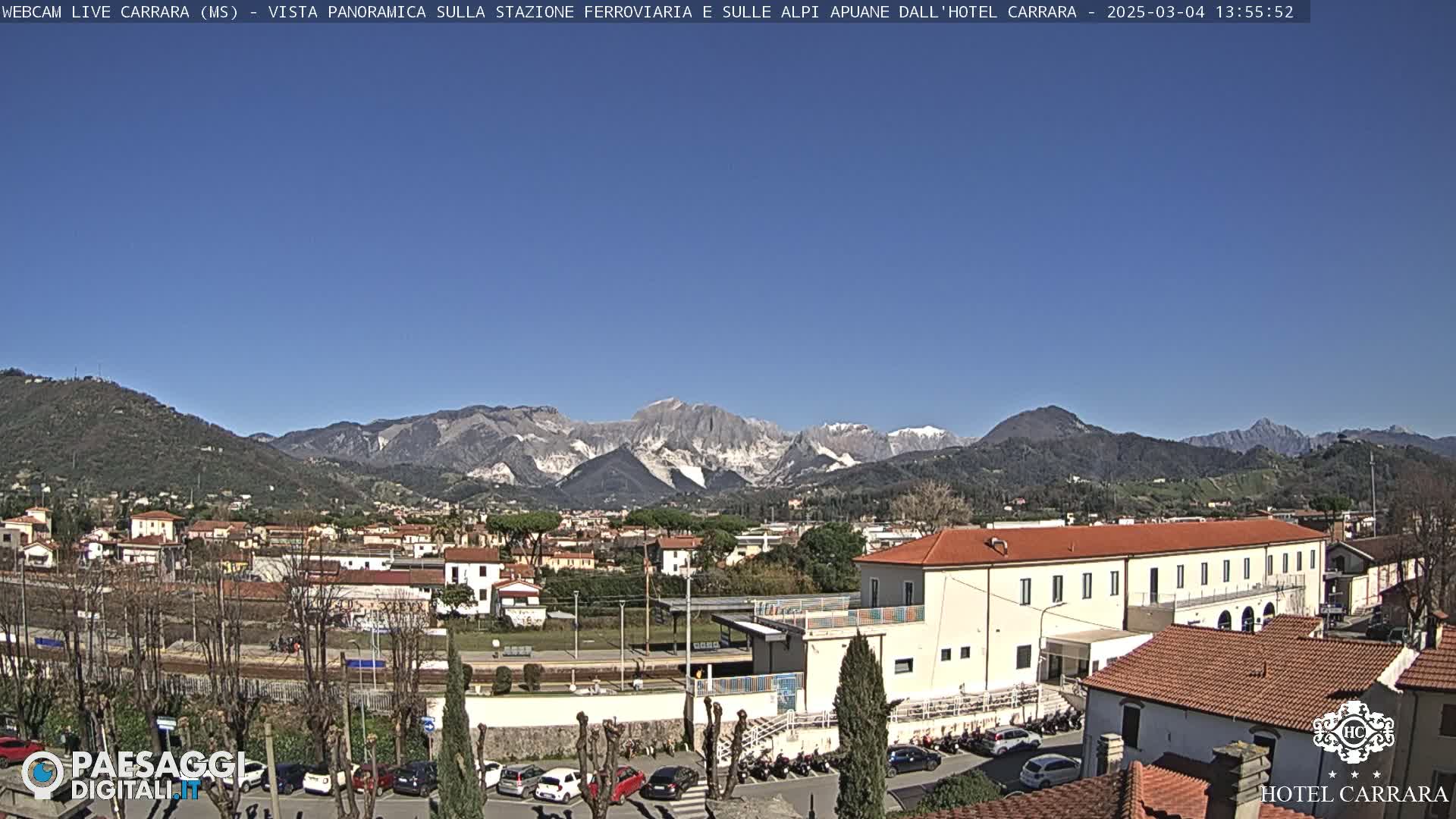 A sunny view of a town nestled in a valley, with snow-capped mountains in the background.