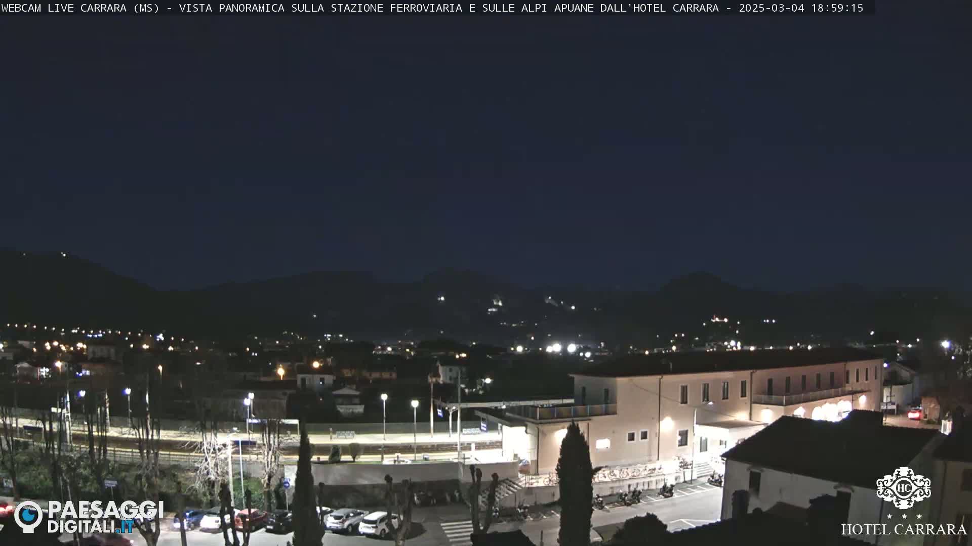 A nighttime panoramic view of a town situated at the foot of dark mountains, showing a train station and a hotel with cars parked in the foreground.