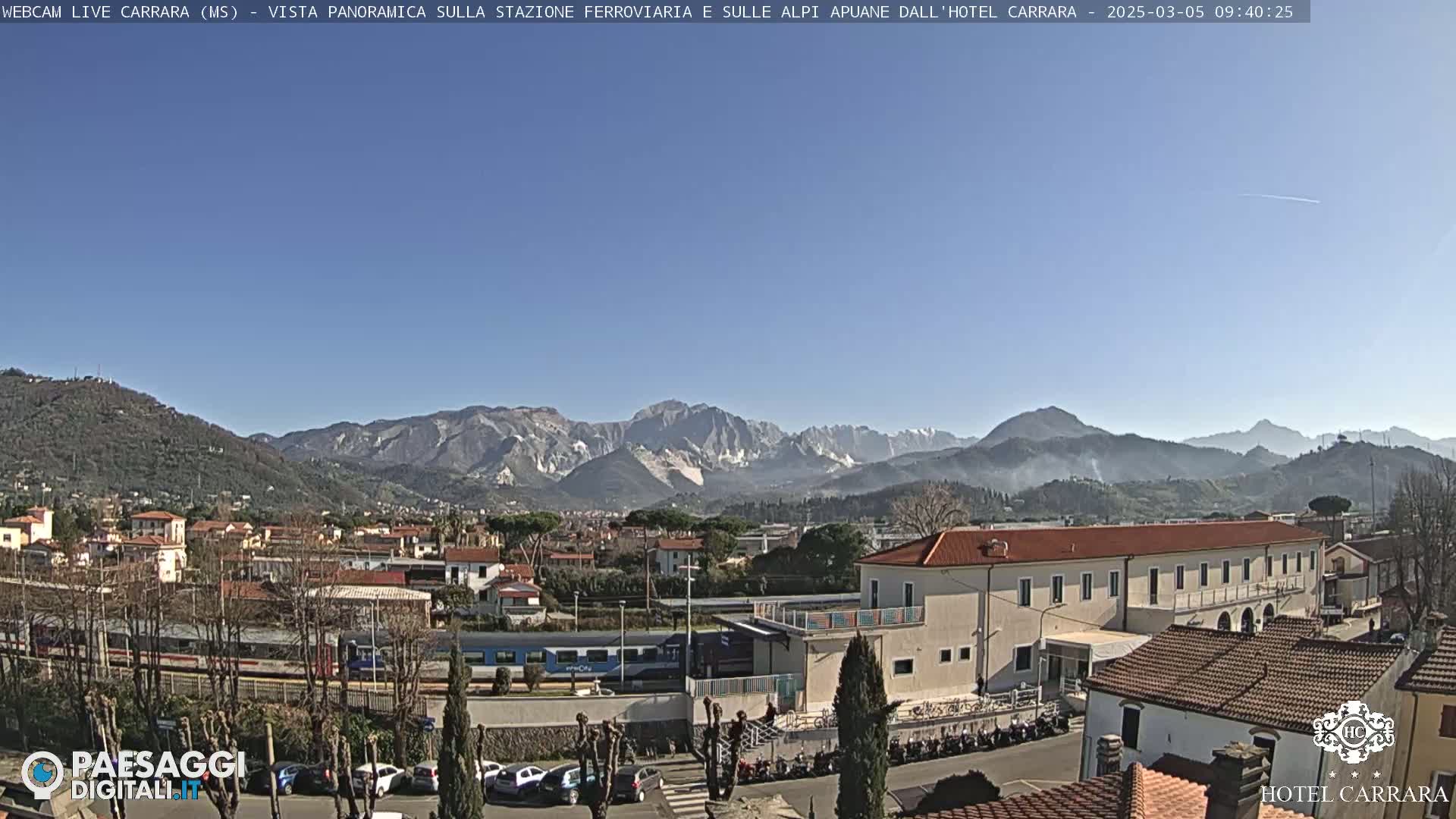 A sunny day shows a train at a station in a town nestled in a valley, with a mountain range in the background.