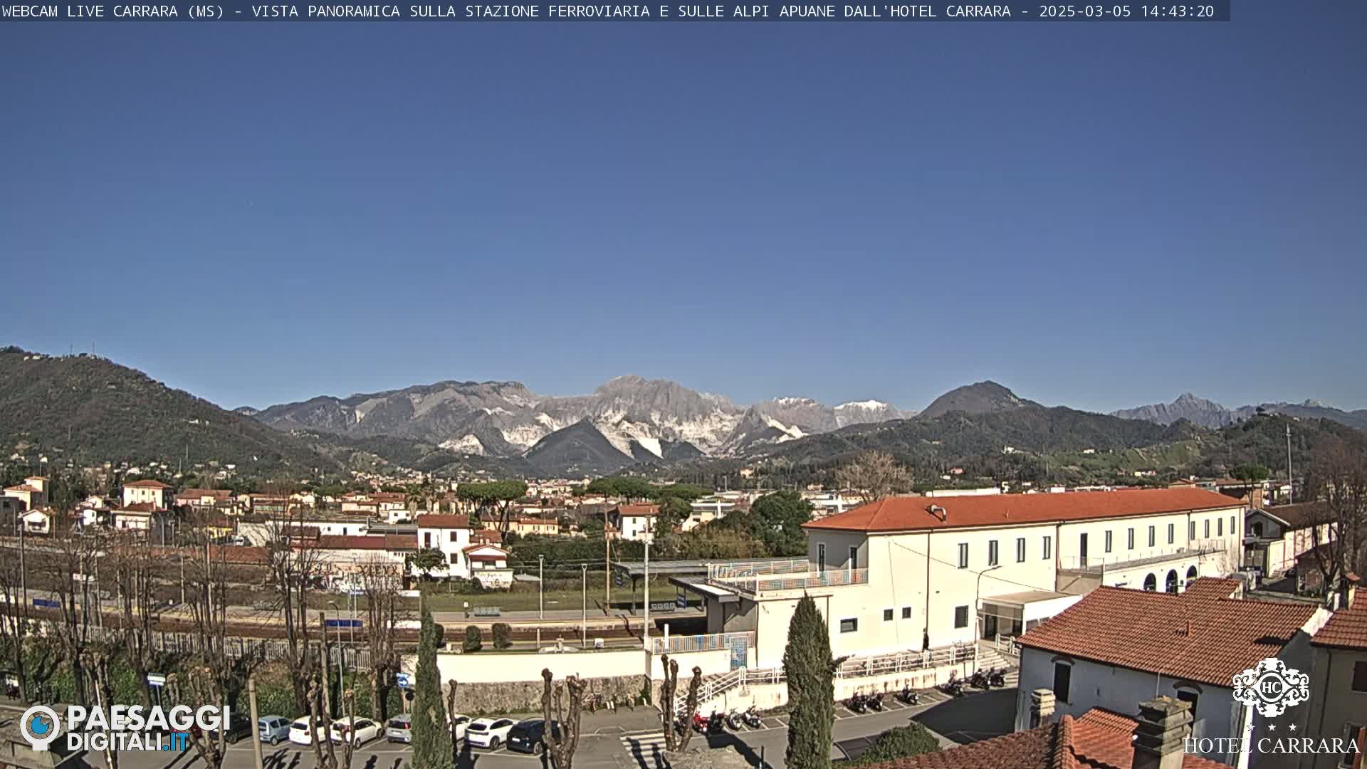 A sunny day reveals a cityscape nestled in a valley, with snow-capped mountains in the background and a train station prominent in the foreground.