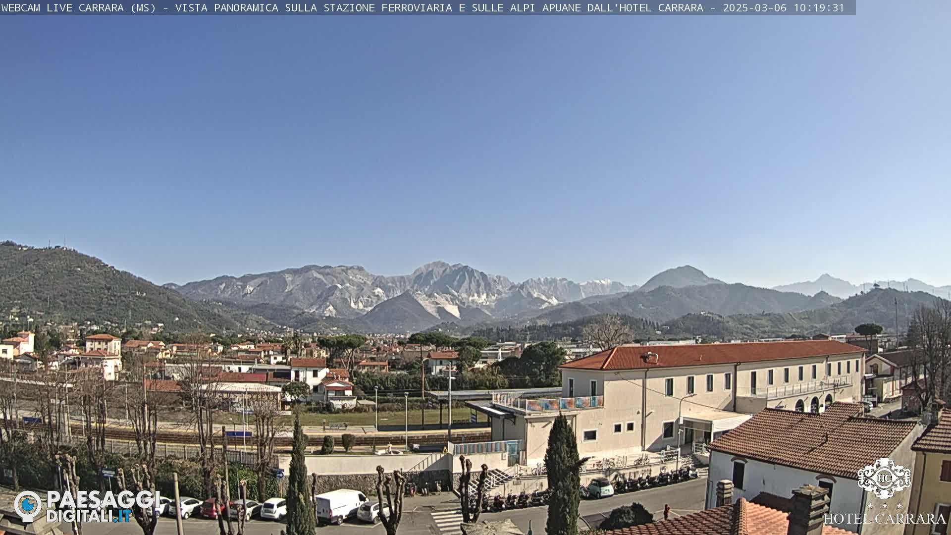 A clear, sunny day reveals a panoramic view of a town nestled in a valley, with a train station and buildings in the foreground and a mountain range in the background.