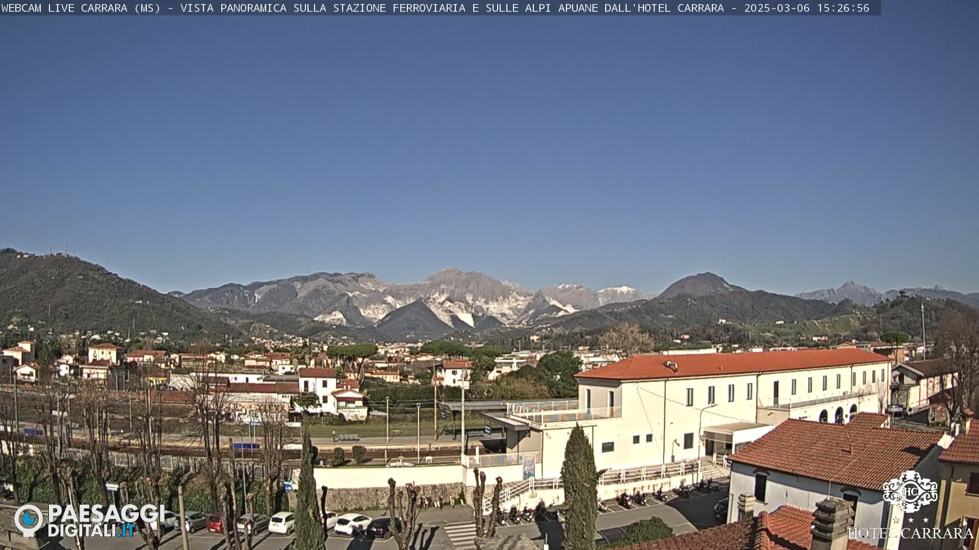 A clear, sunny day reveals a panoramic view of a town nestled in a valley, with snow-capped mountains in the background and a train station in the foreground.