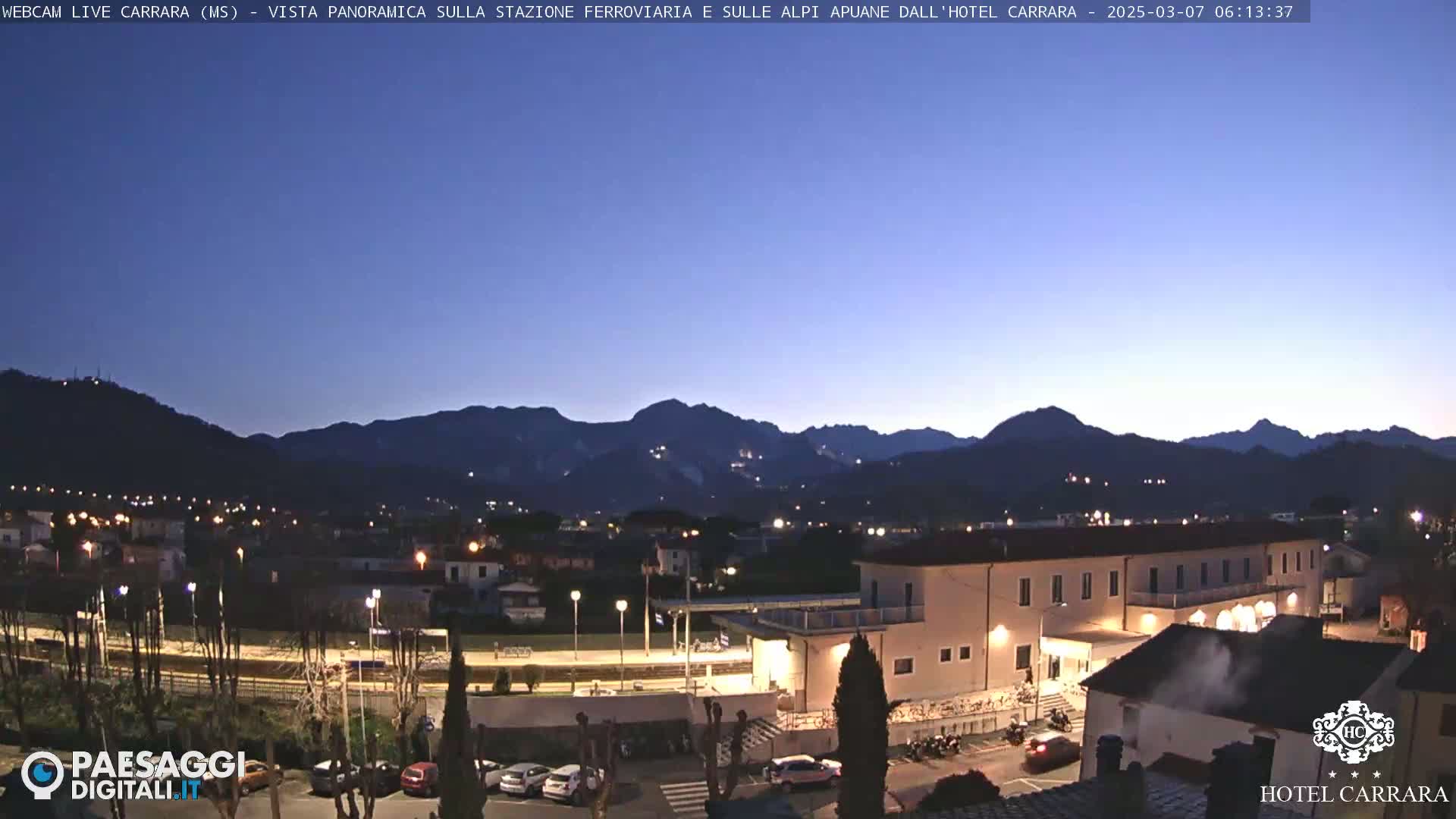 A twilight view of a town nestled in a valley, with mountains in the background and a building illuminated by lights.