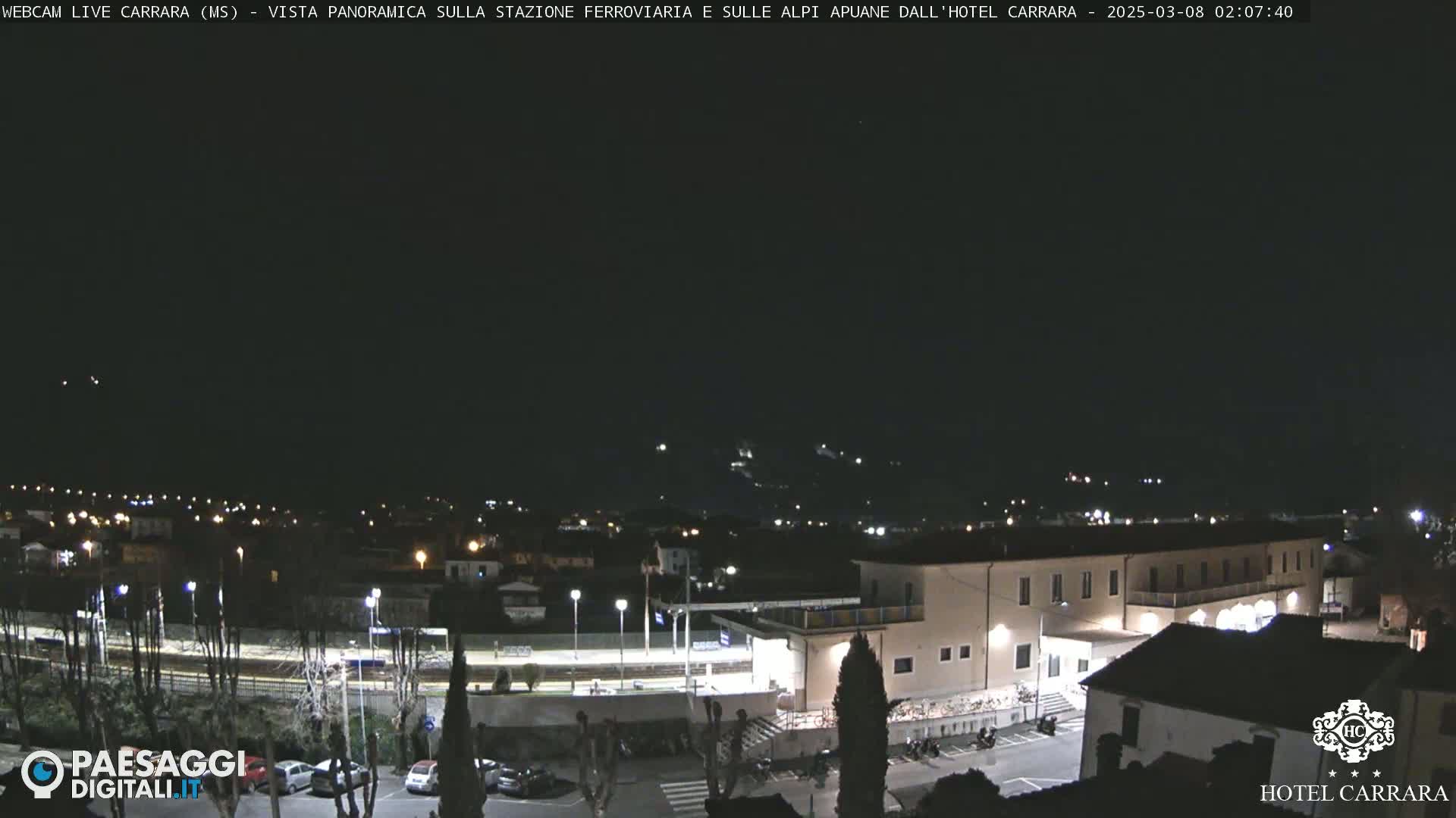 A nighttime panoramic view of a town's outskirts, featuring a train station and a large building, under a clear, dark sky.