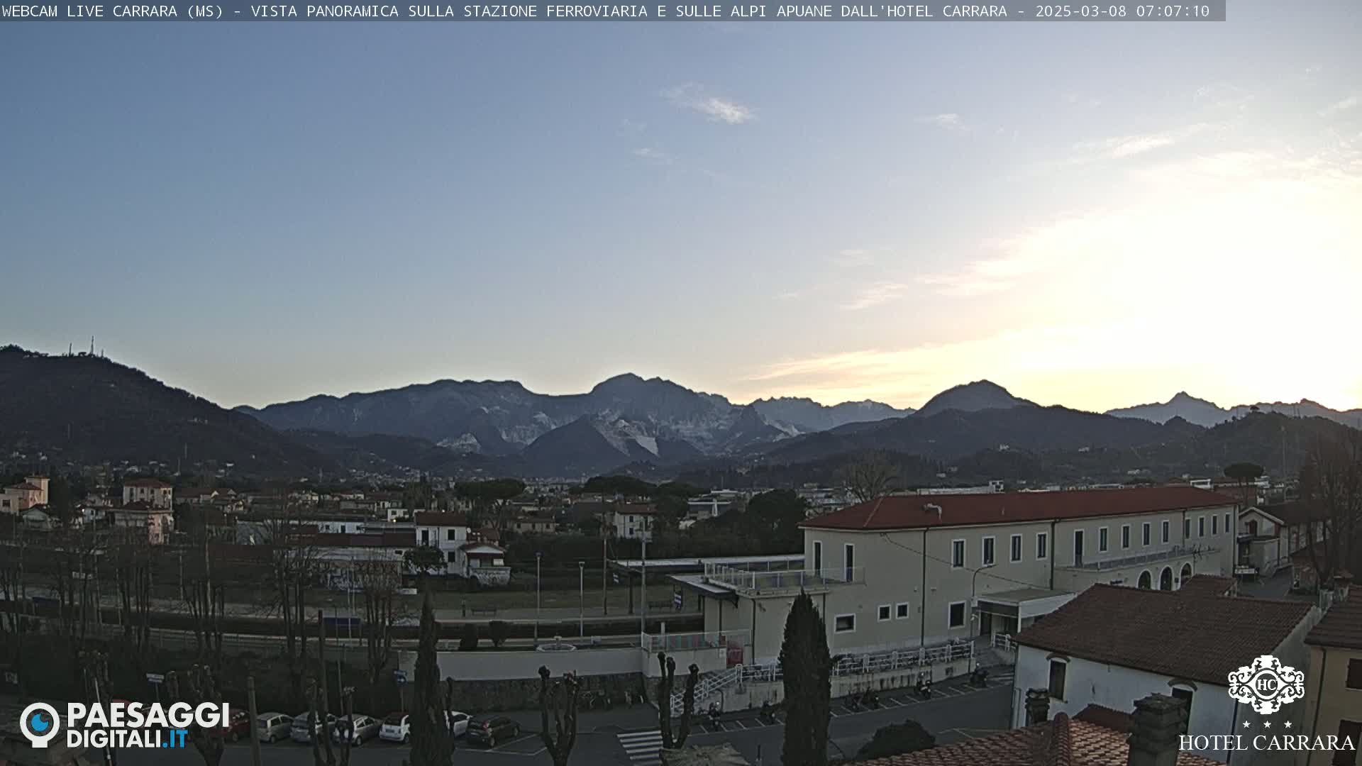 A clear sunrise illuminates a snow-capped mountain range behind a town with buildings and a train station.