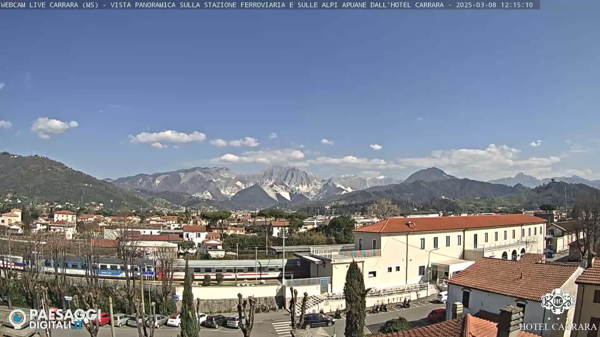 A sunny day reveals a train station and surrounding buildings nestled in a valley, with a mountain range in the background under a partly cloudy sky.