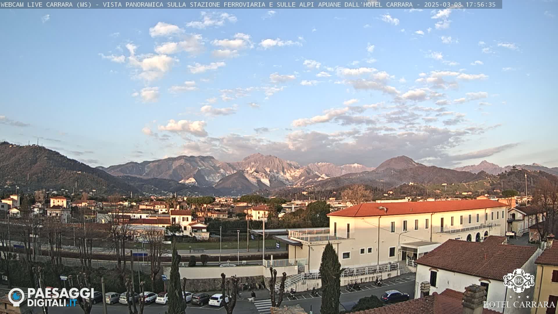 A partly cloudy day reveals a panoramic view of a town nestled amongst mountains, with a large building in the foreground.