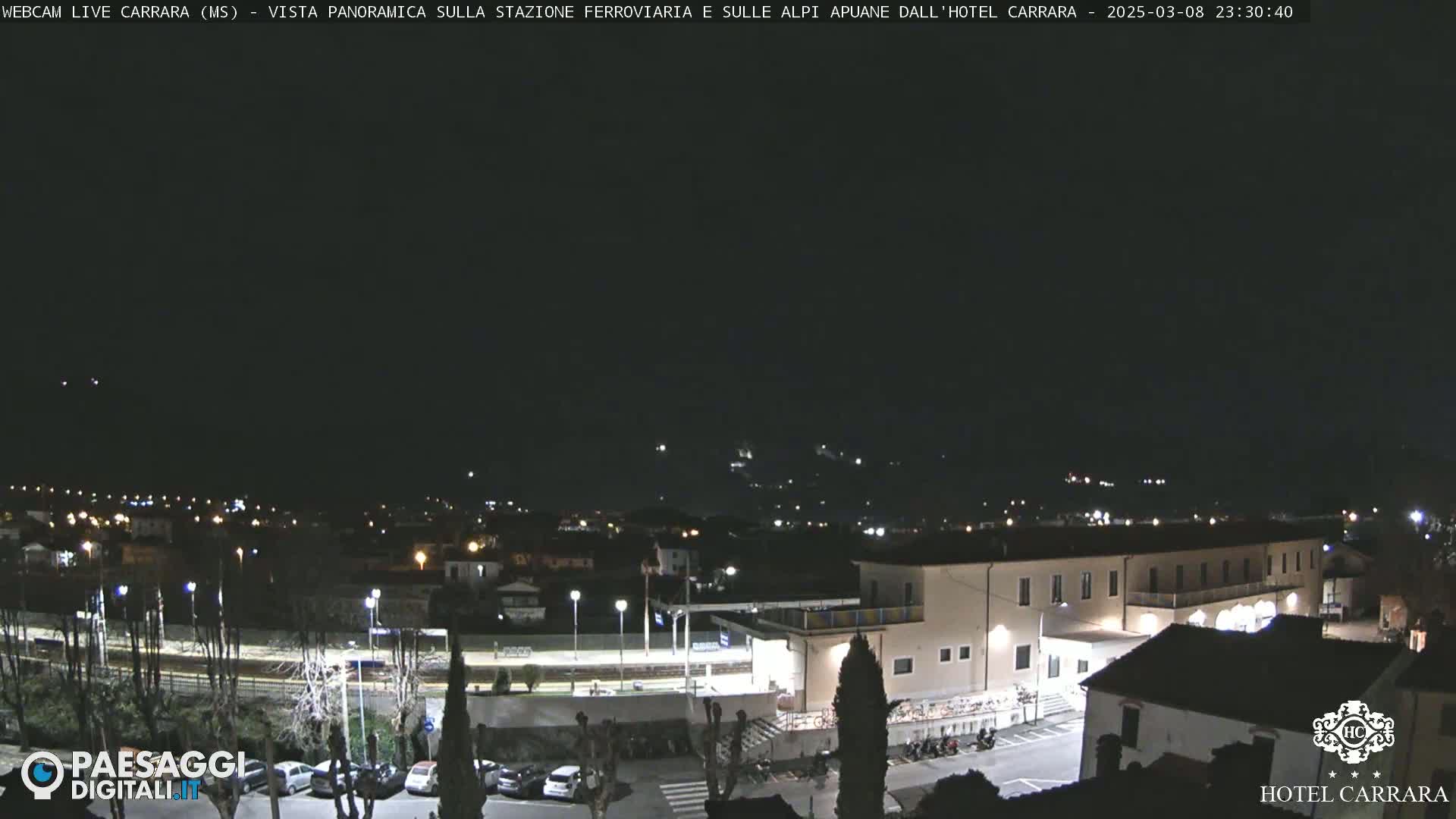 A nighttime long shot of a town with a train station and a large building, under a clear, dark sky.
