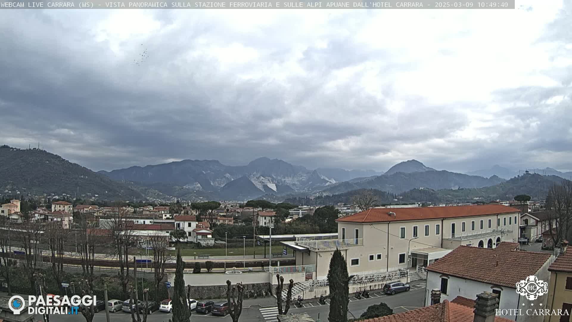 A panoramic view shows a town nestled in a valley, with mountains in the background under a cloudy sky.