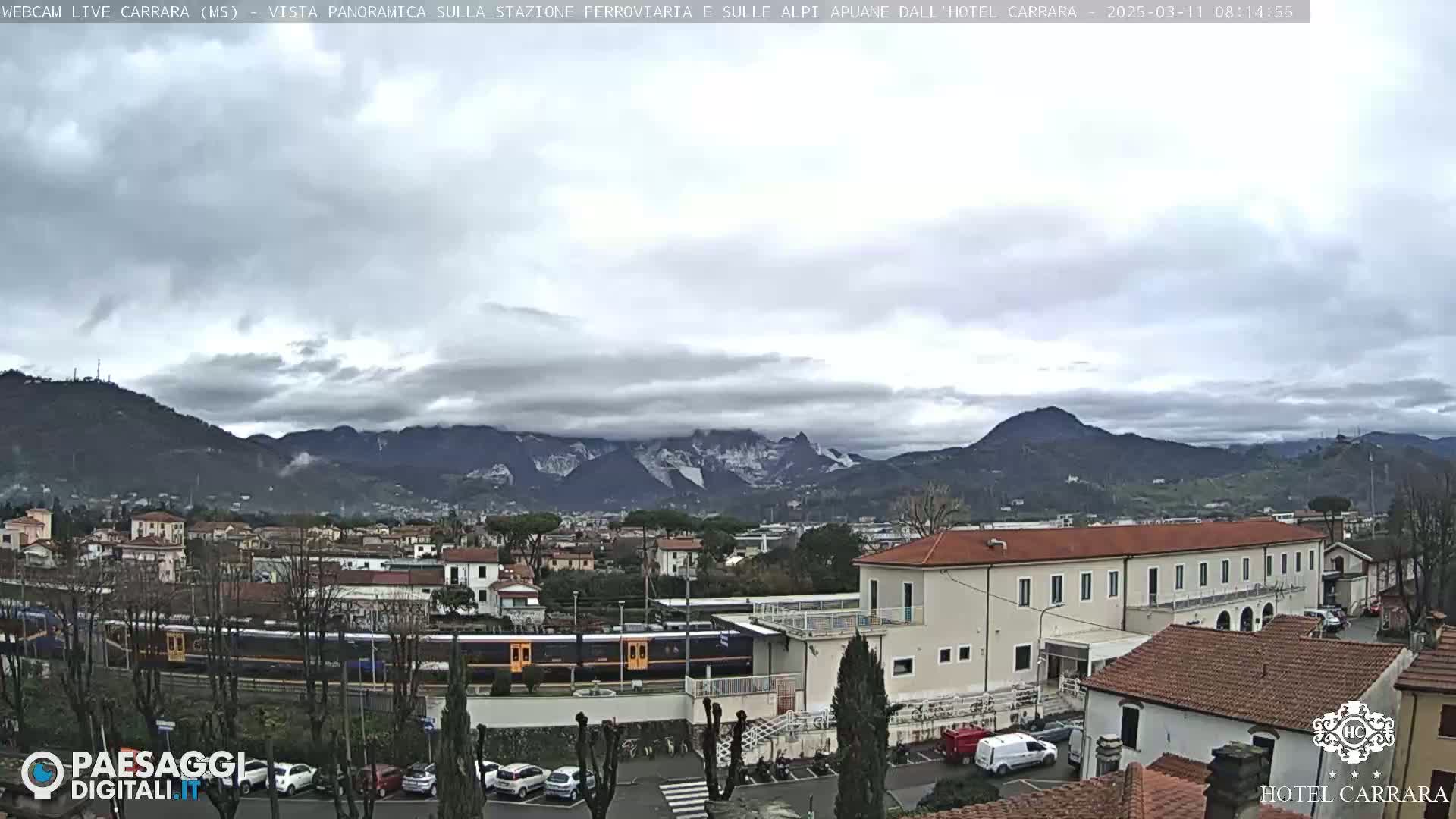 A long train sits in a station next to a long building, all of which are nestled in a valley under a mostly cloudy sky and snow-capped mountains in the distance.