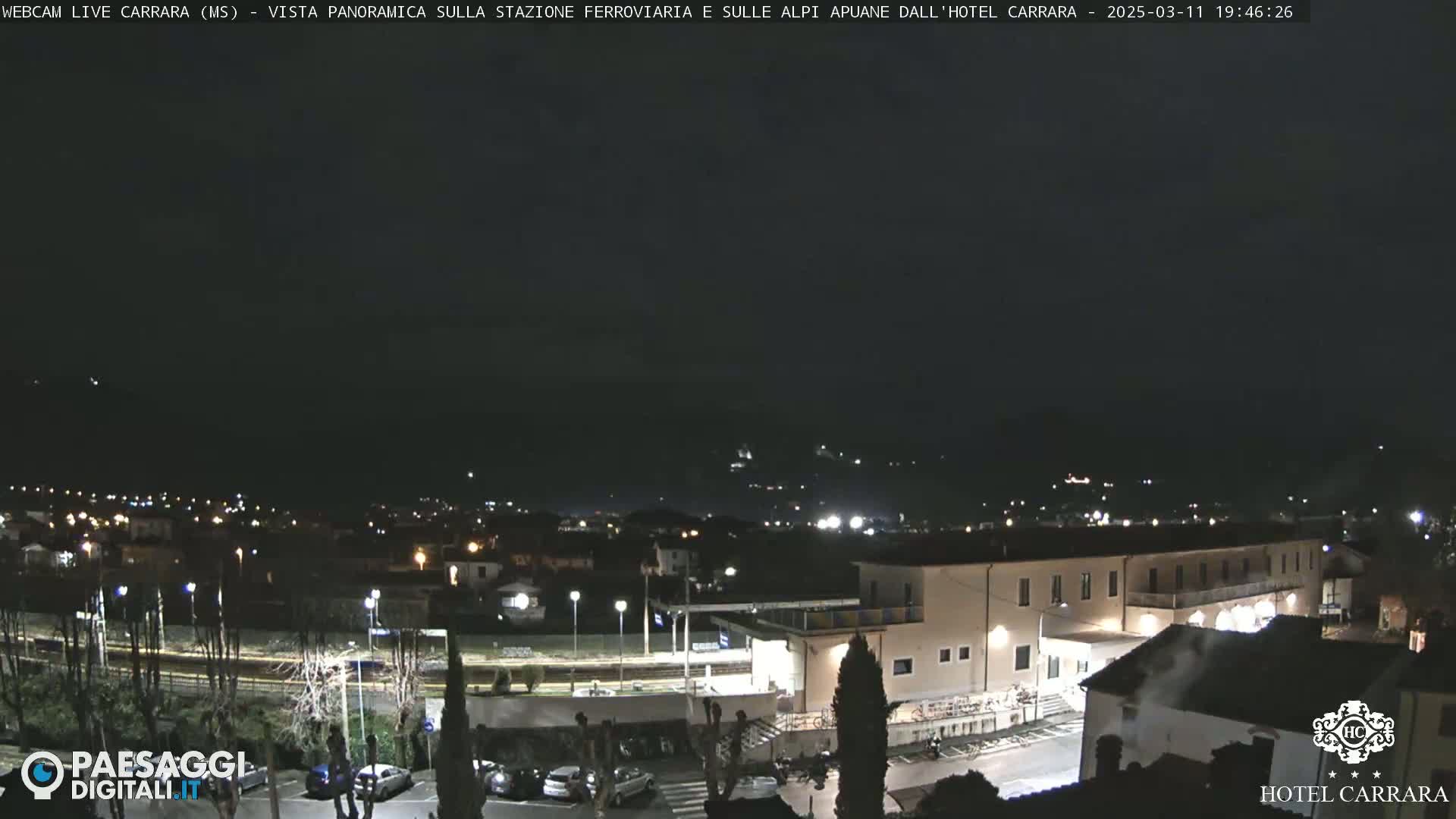 A nighttime, panoramic view of a town with a train station and a large building, possibly a hotel, under a dark, clear sky.