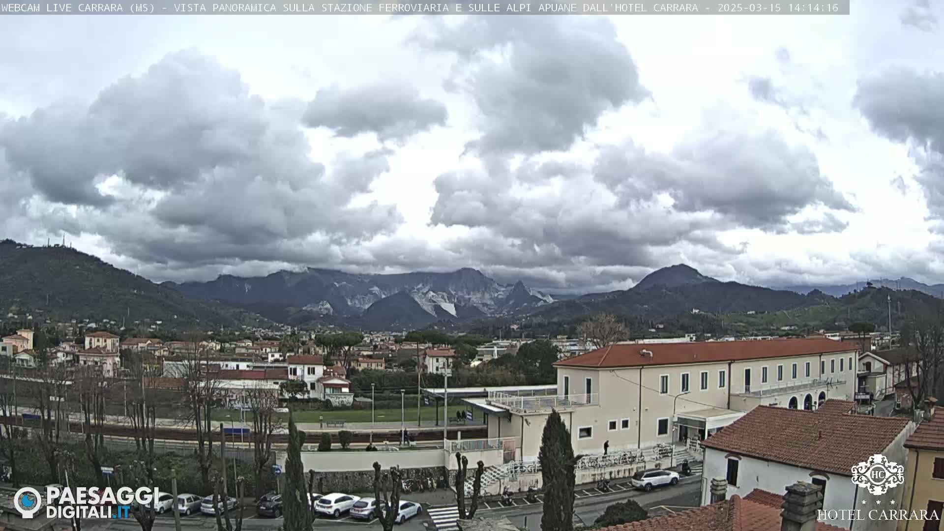 A panoramic view of a town nestled in a valley, with a cloudy sky and mountains in the background.