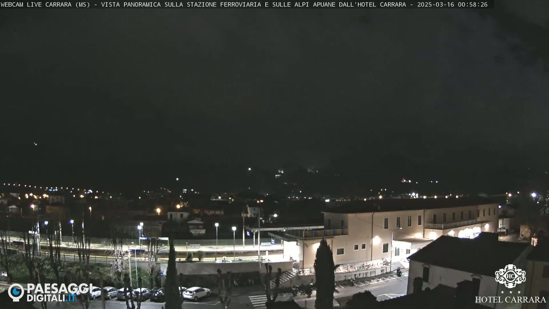 A nighttime long shot of a town with a train station and hotel, under a dark, overcast sky.