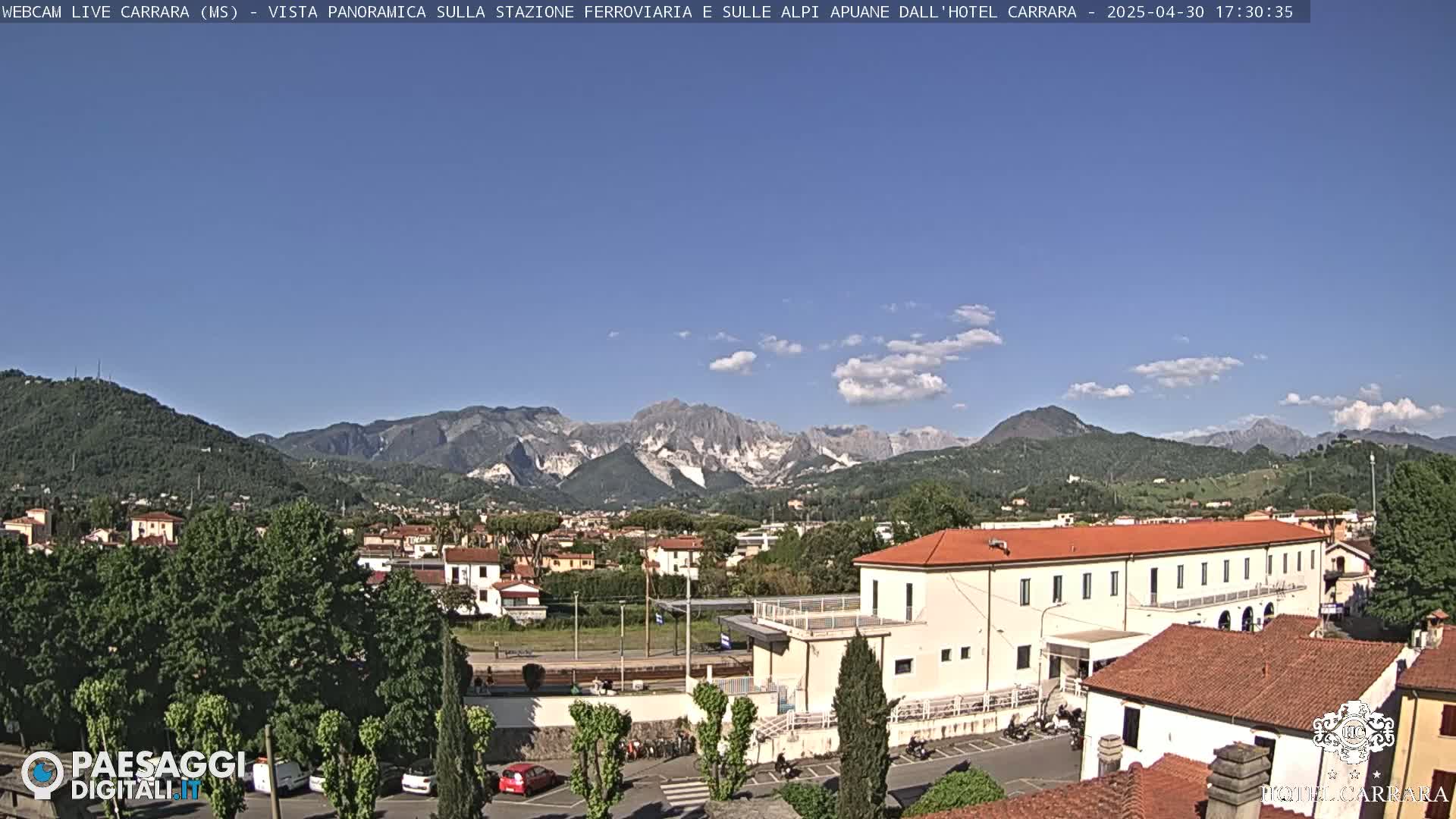 A clear, sunny day reveals a panoramic view of a town nestled in a valley, with a large building in the foreground and mountains in the background.