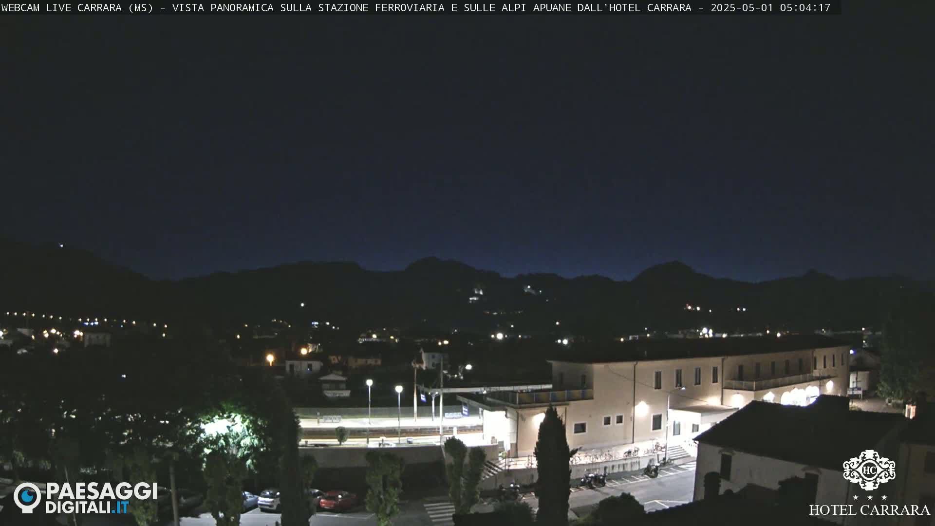A nighttime view shows a town nestled among dark mountains under a clear, dark sky, with a large building illuminated in the foreground.