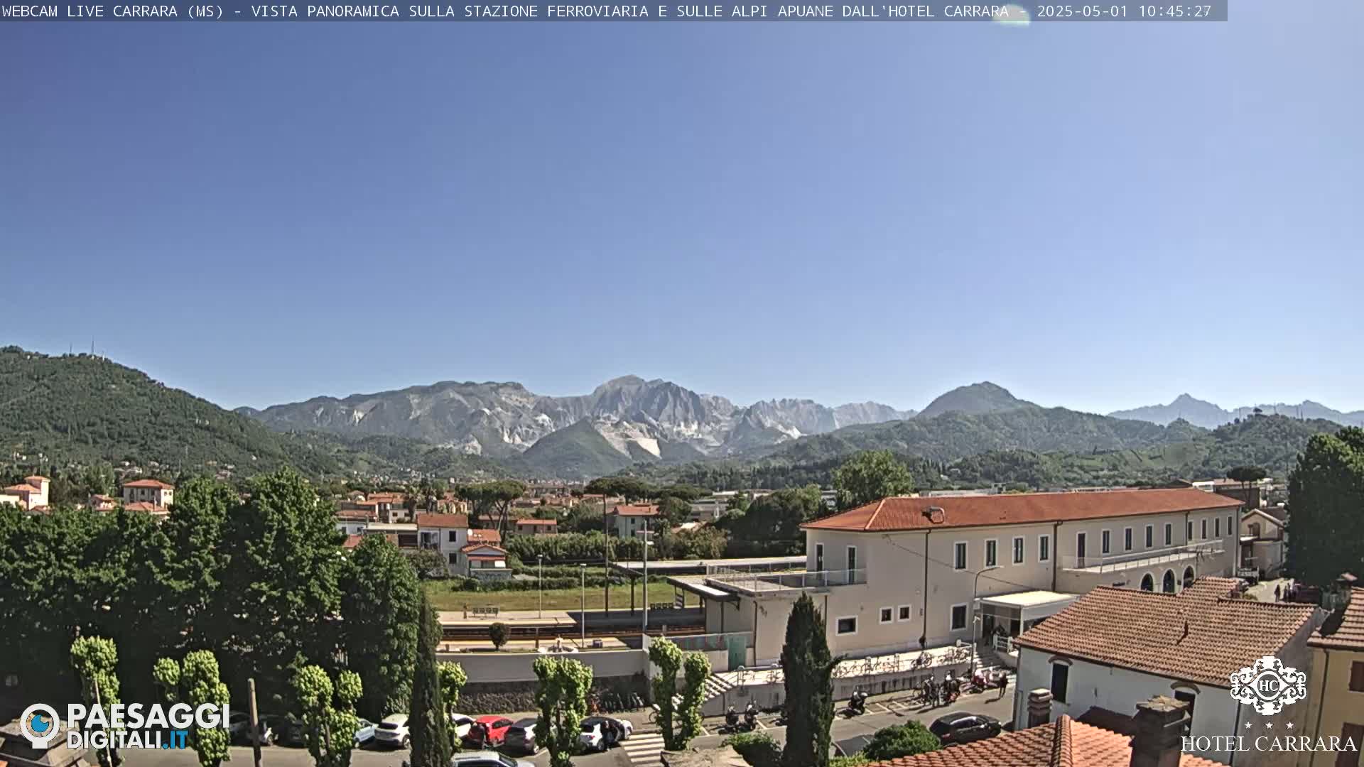 A sunny view shows a town nestled between green hills and a mountain range in the distance, with a train station and buildings in the foreground.