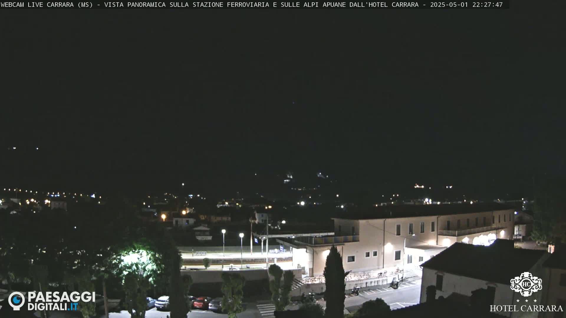 A nighttime, panoramic view of a town's outskirts, featuring a brightly lit hotel and a train station, under a clear, dark sky.