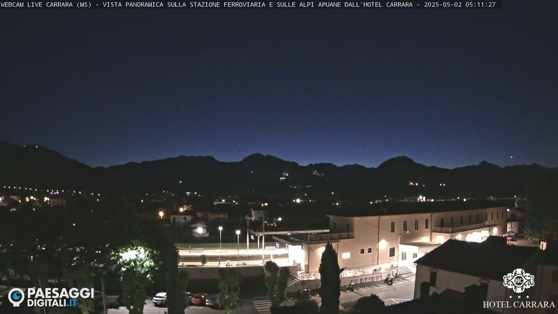 A nighttime view of a town nestled in a valley, with mountains silhouetted against a dark blue sky.