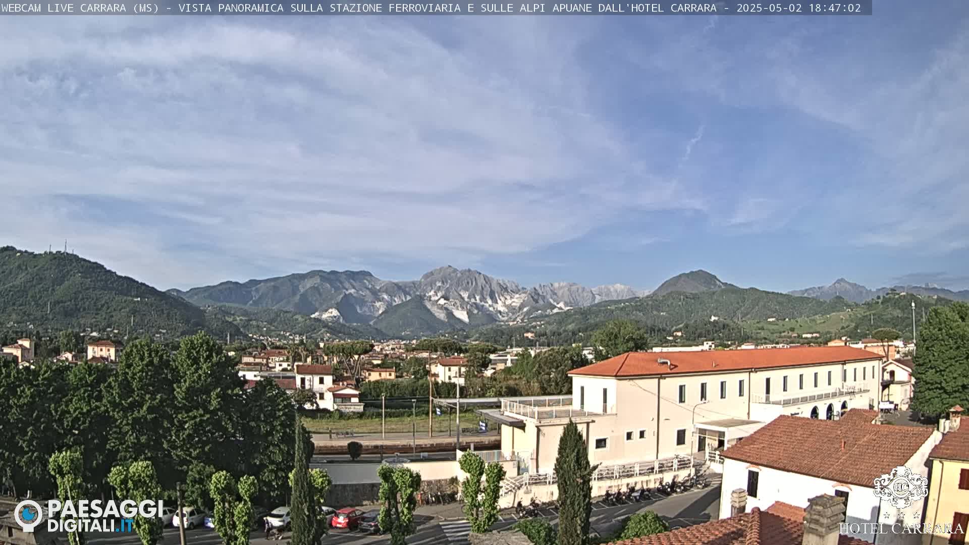 A partly cloudy daytime view shows a town nestled in a valley between mountains.