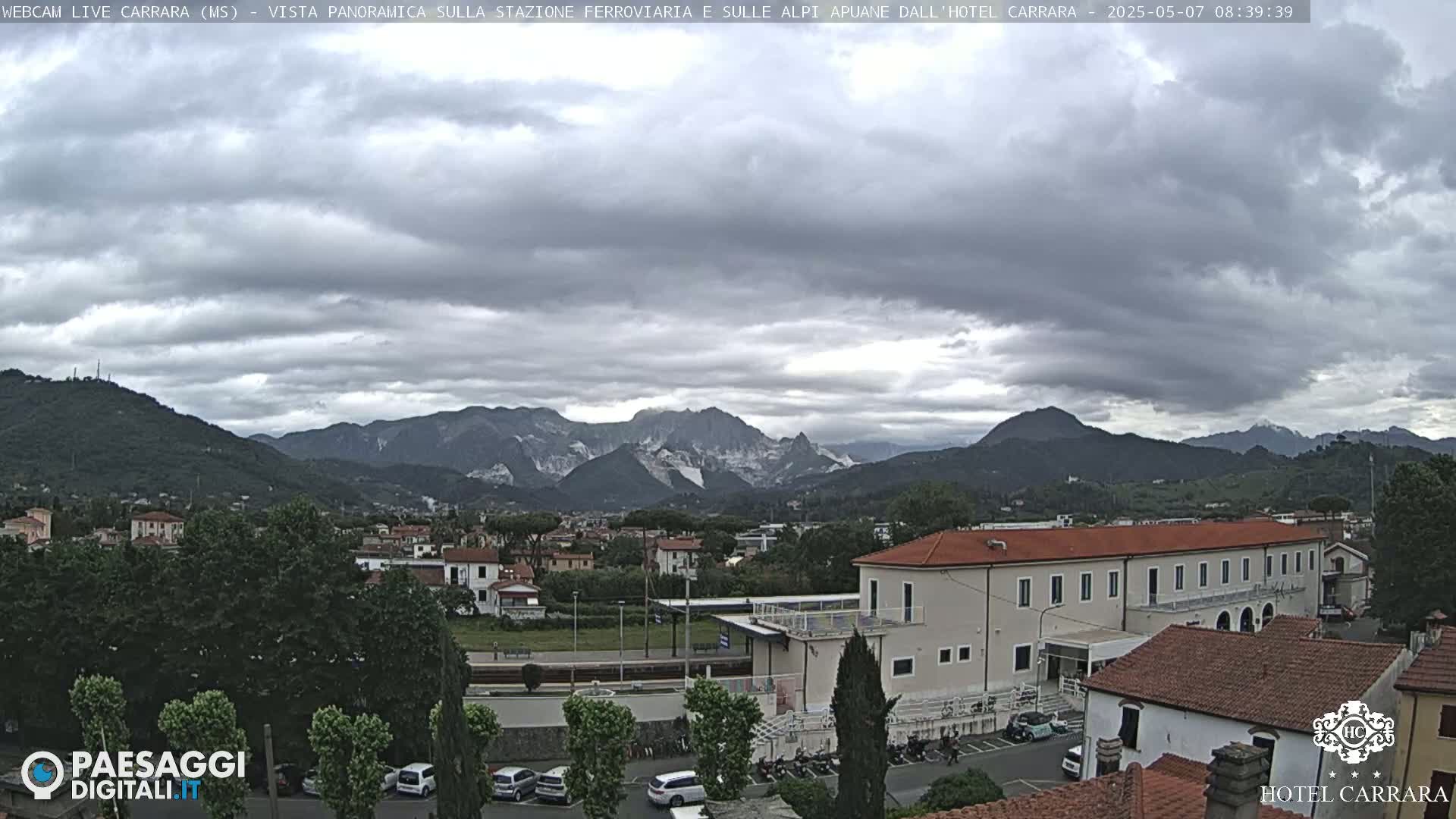 A long shot of a town nestled in a valley, overlooked by mountains under a cloudy sky.