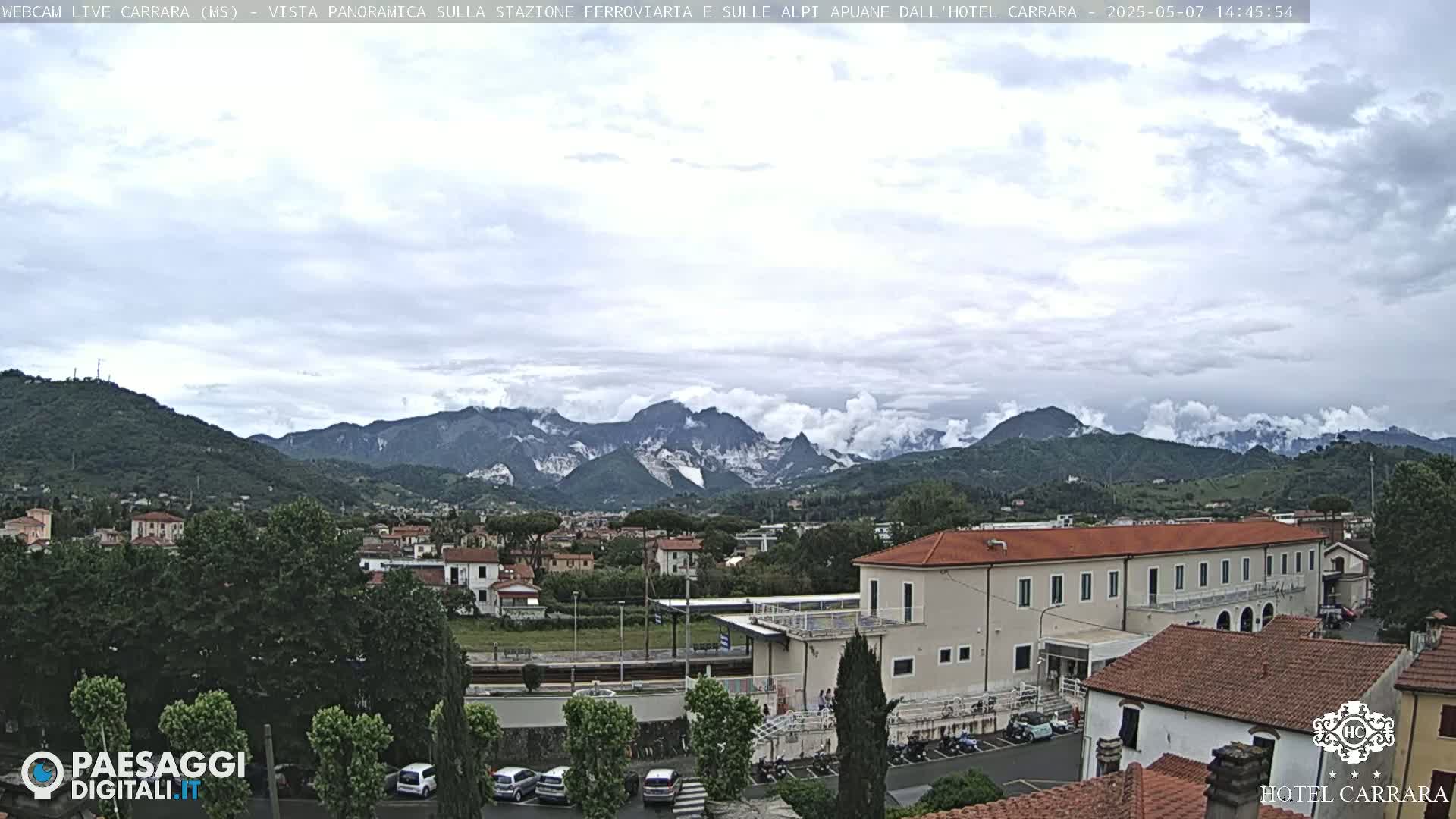 A cloudy day reveals a panoramic view of a town nestled in a valley, with mountains in the background and a train station visible in the foreground.