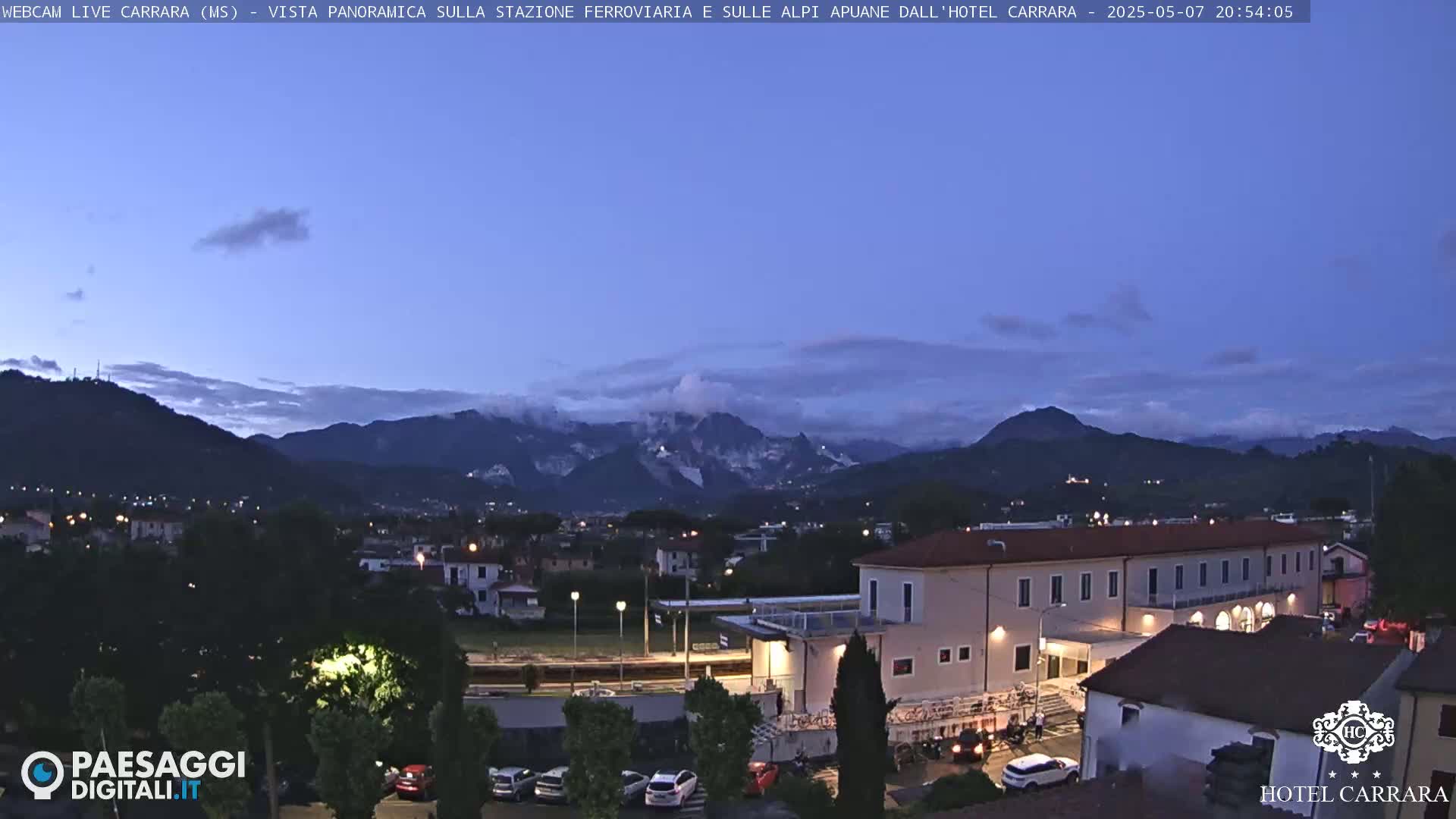 A twilight view of a town nestled in a valley, with mountains in the background under a mostly clear, dusky sky.