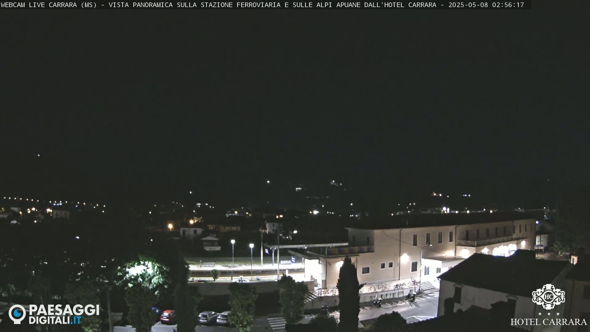 A nighttime, panoramic view of a town with a train station and a large building, possibly a hotel, under a clear, dark sky.