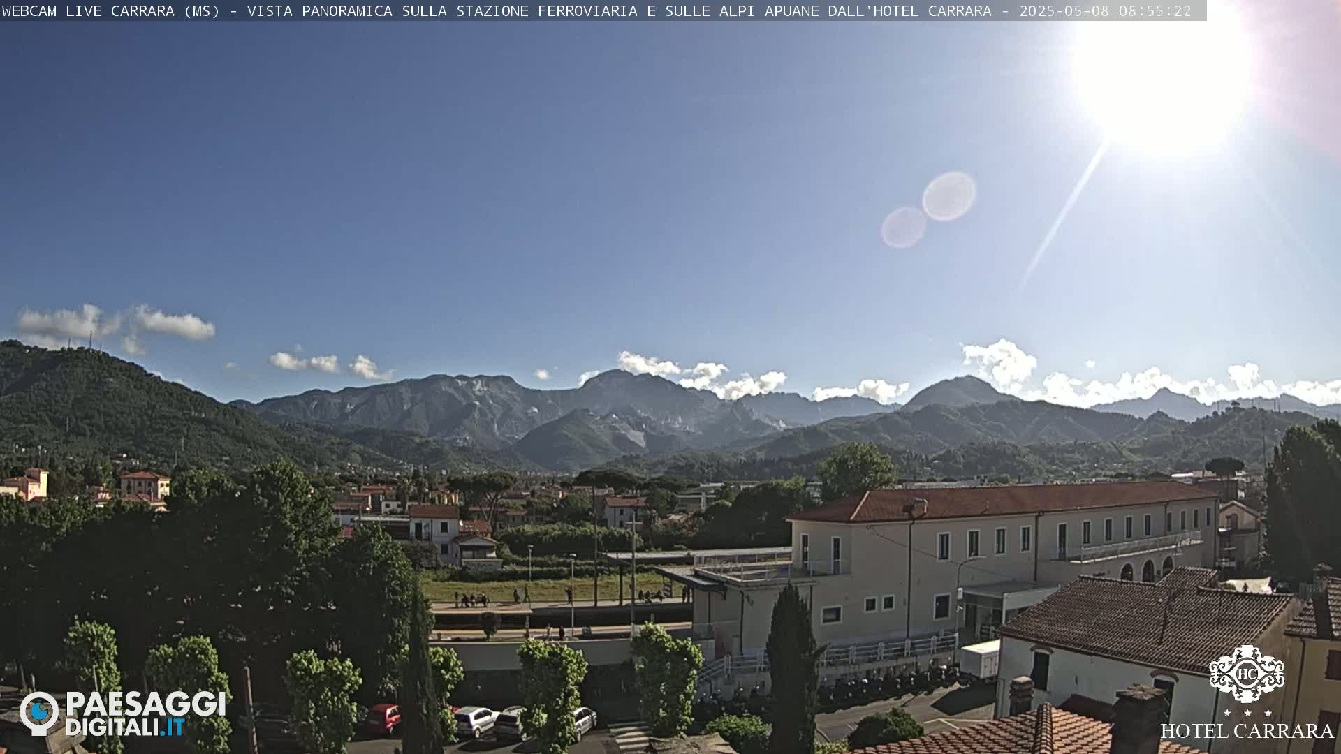 A sunny day reveals a picturesque view of a town nestled at the foot of snow-capped mountains, with a train station and buildings visible in the foreground.