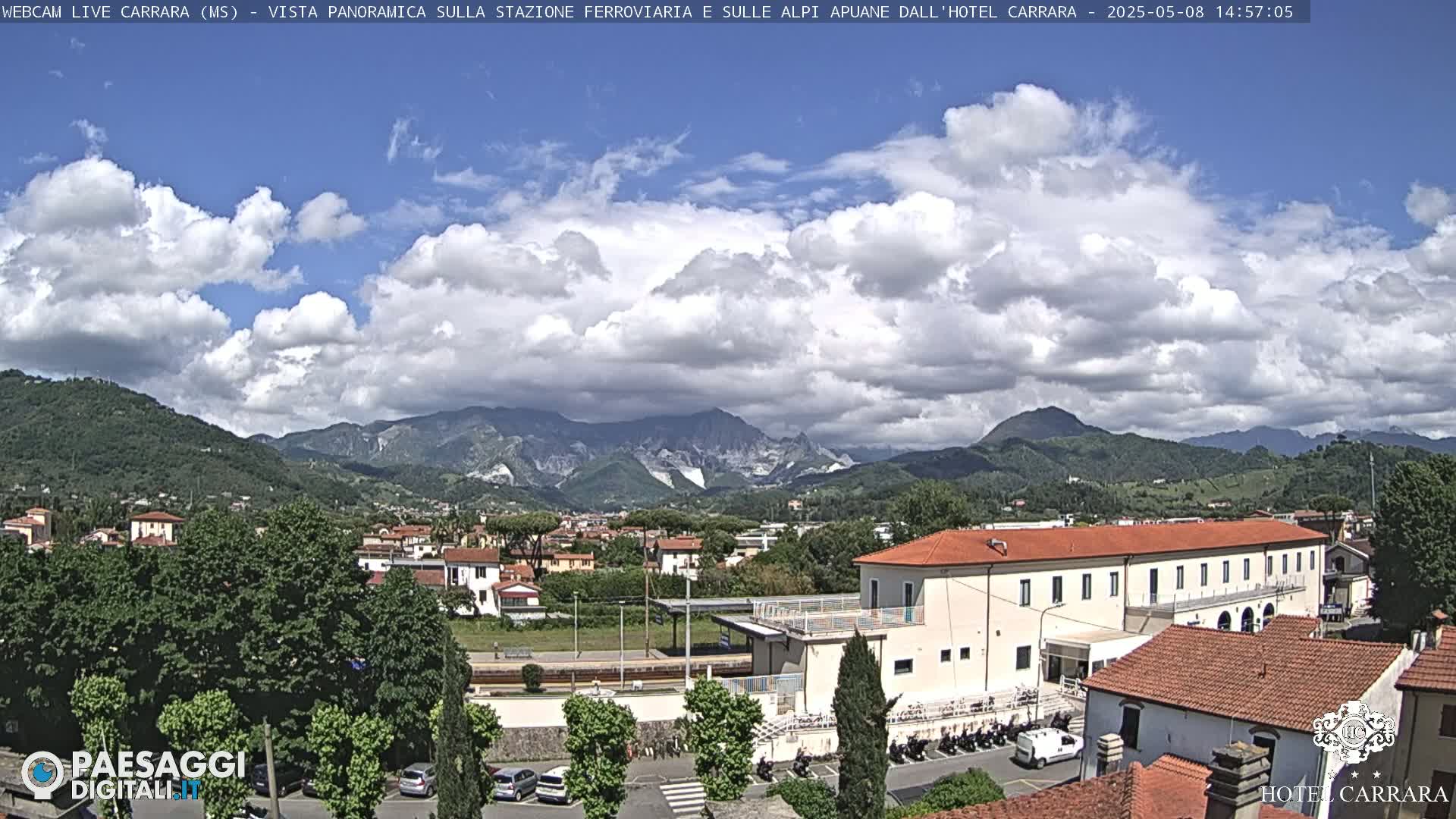 A partly cloudy day reveals a panoramic view of a town nestled in a valley, with mountains in the background and a train station visible in the foreground.