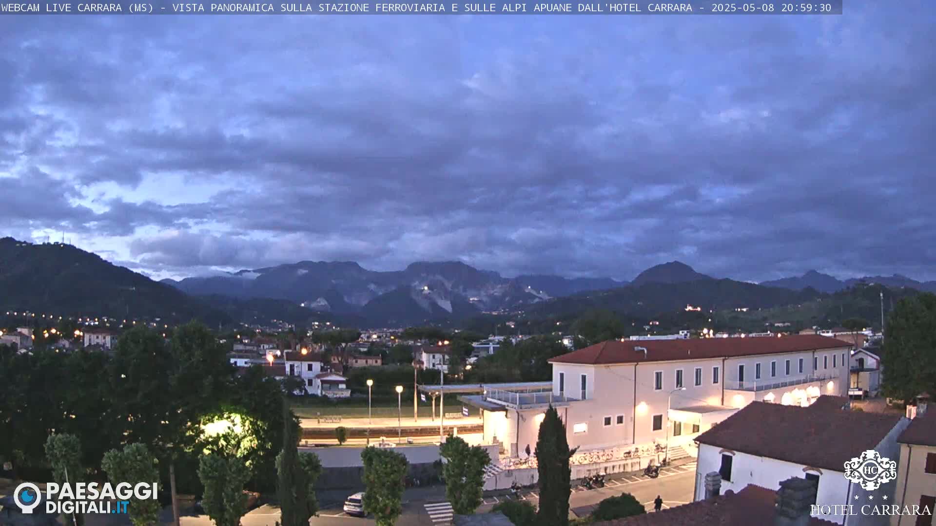 A twilight view of a town nestled in a valley between mountains under a partly cloudy sky.