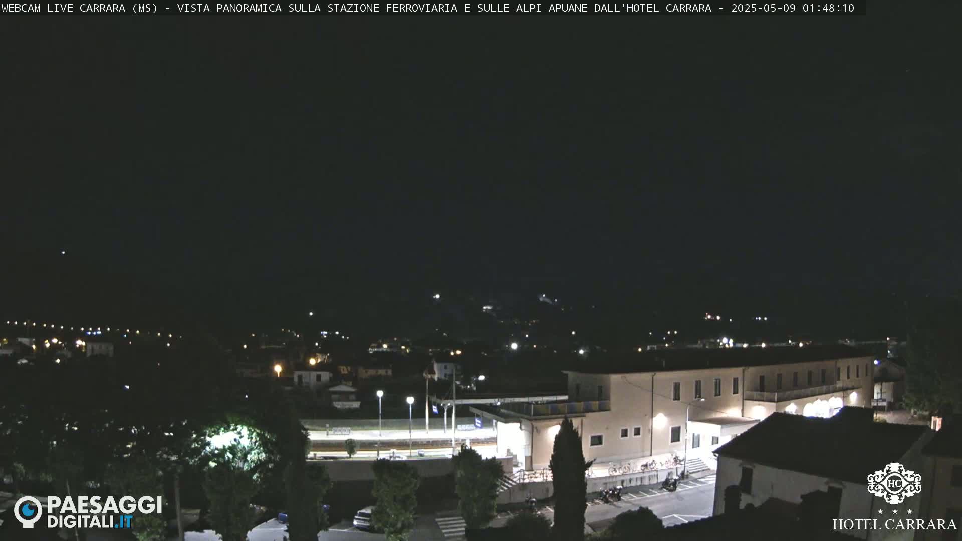 A nighttime, panoramic view of a town, featuring a large building illuminated, and a train station, under a clear, dark sky.