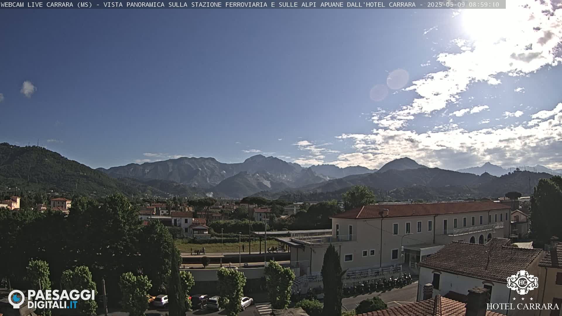 A partly cloudy sky overlooks a town nestled in a valley, with mountains in the background.