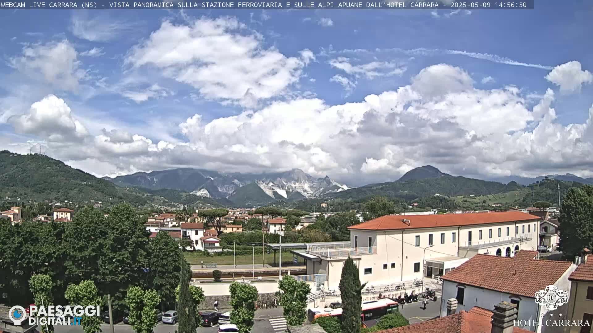 A partly cloudy day reveals a panoramic view of a town nestled in a valley, with mountains in the background and a train station visible in the foreground.