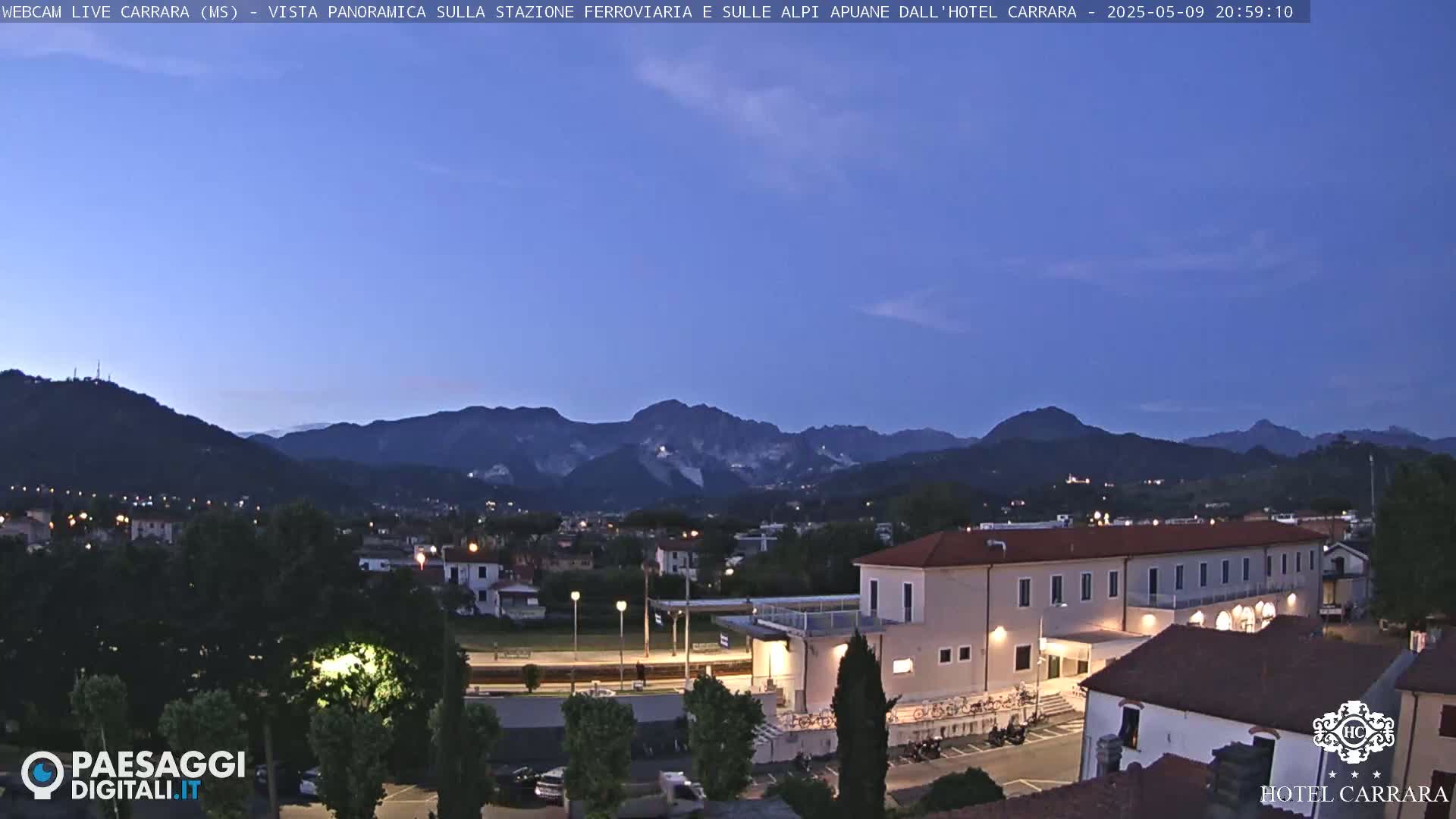 A twilight view shows a town nestled in a valley, with mountains in the background under a clear, dark blue sky.