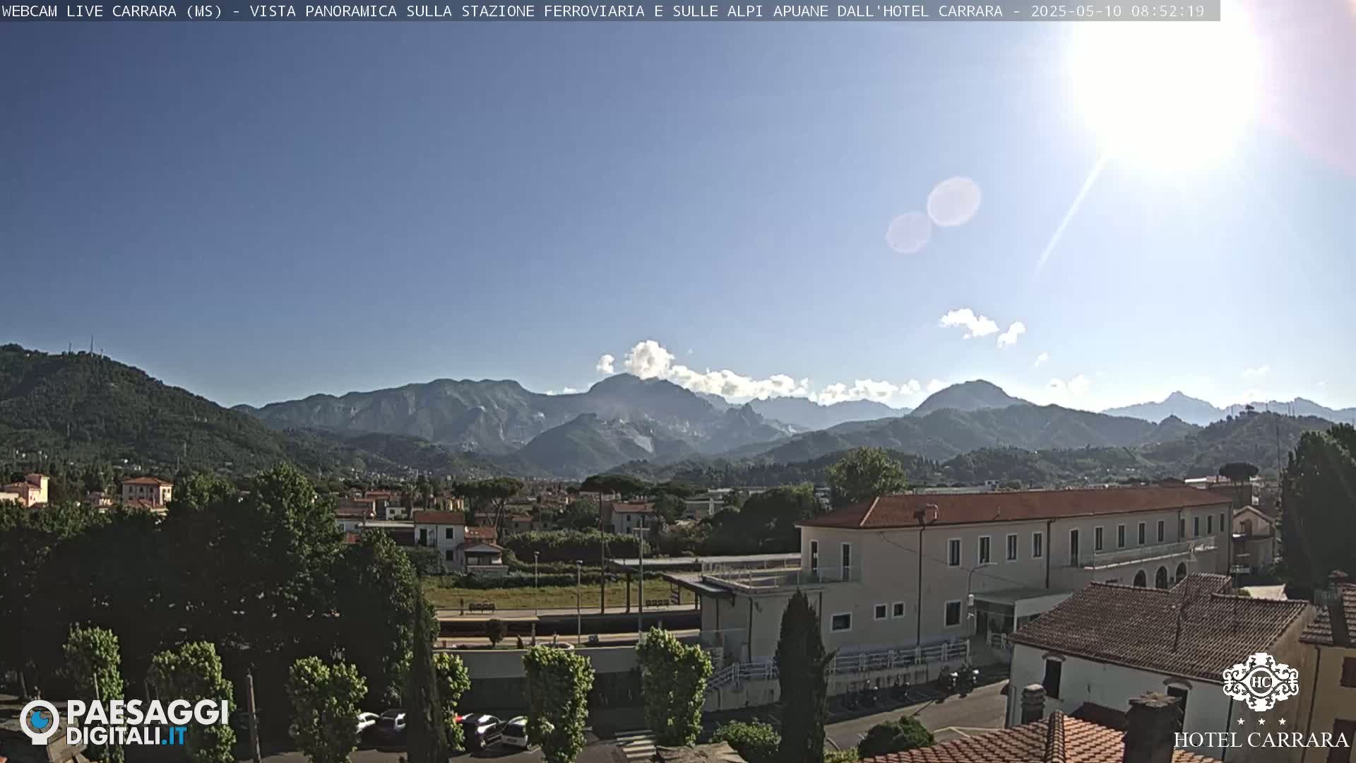 A sunny day reveals a panoramic view of a town nestled amongst lush green hills and a mountain range in the background.