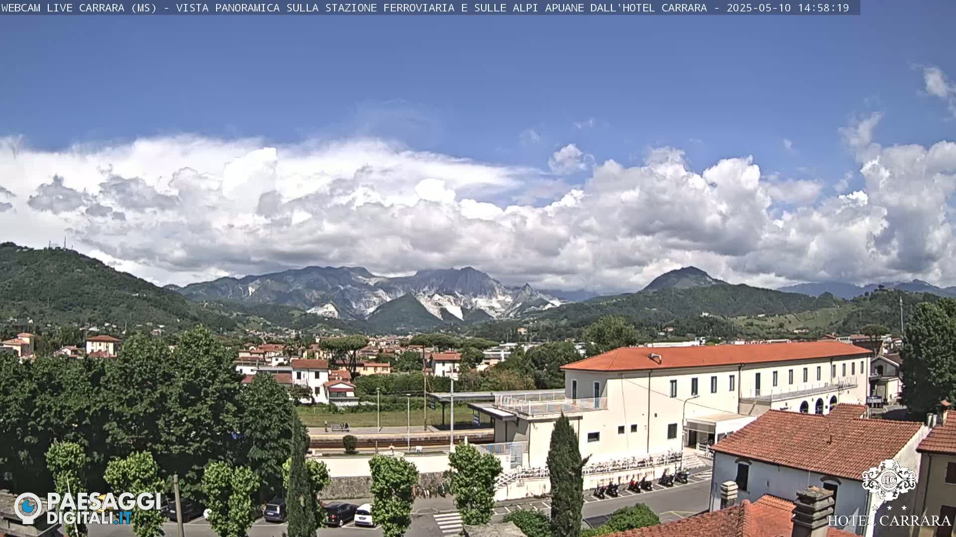 A partly cloudy day reveals a panoramic view of a town nestled among green hills and mountains, with a train station and buildings visible in the foreground.