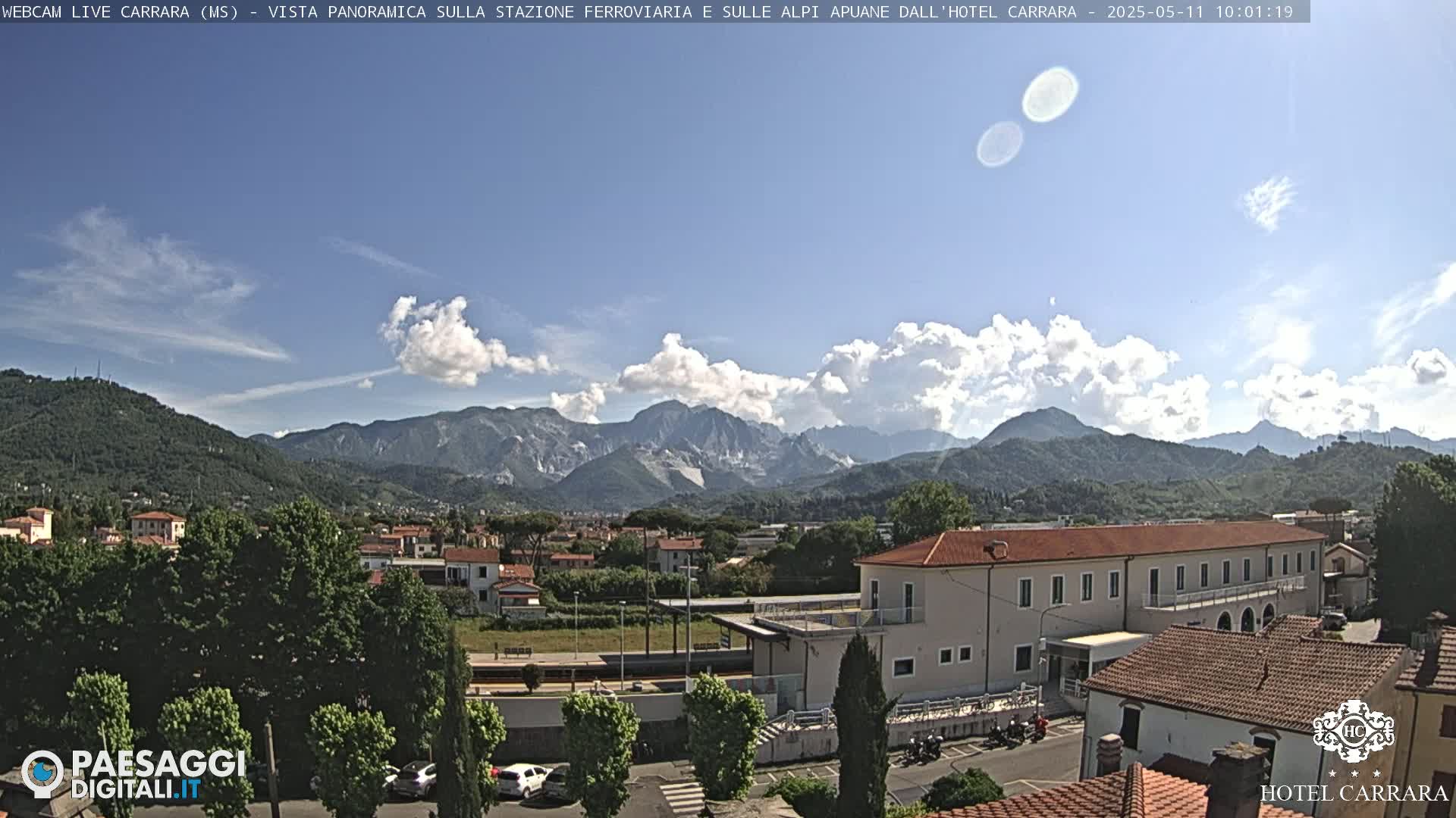 A partly cloudy day reveals a panoramic view of a town nestled in a valley, with mountains in the background and a train station visible in the foreground.