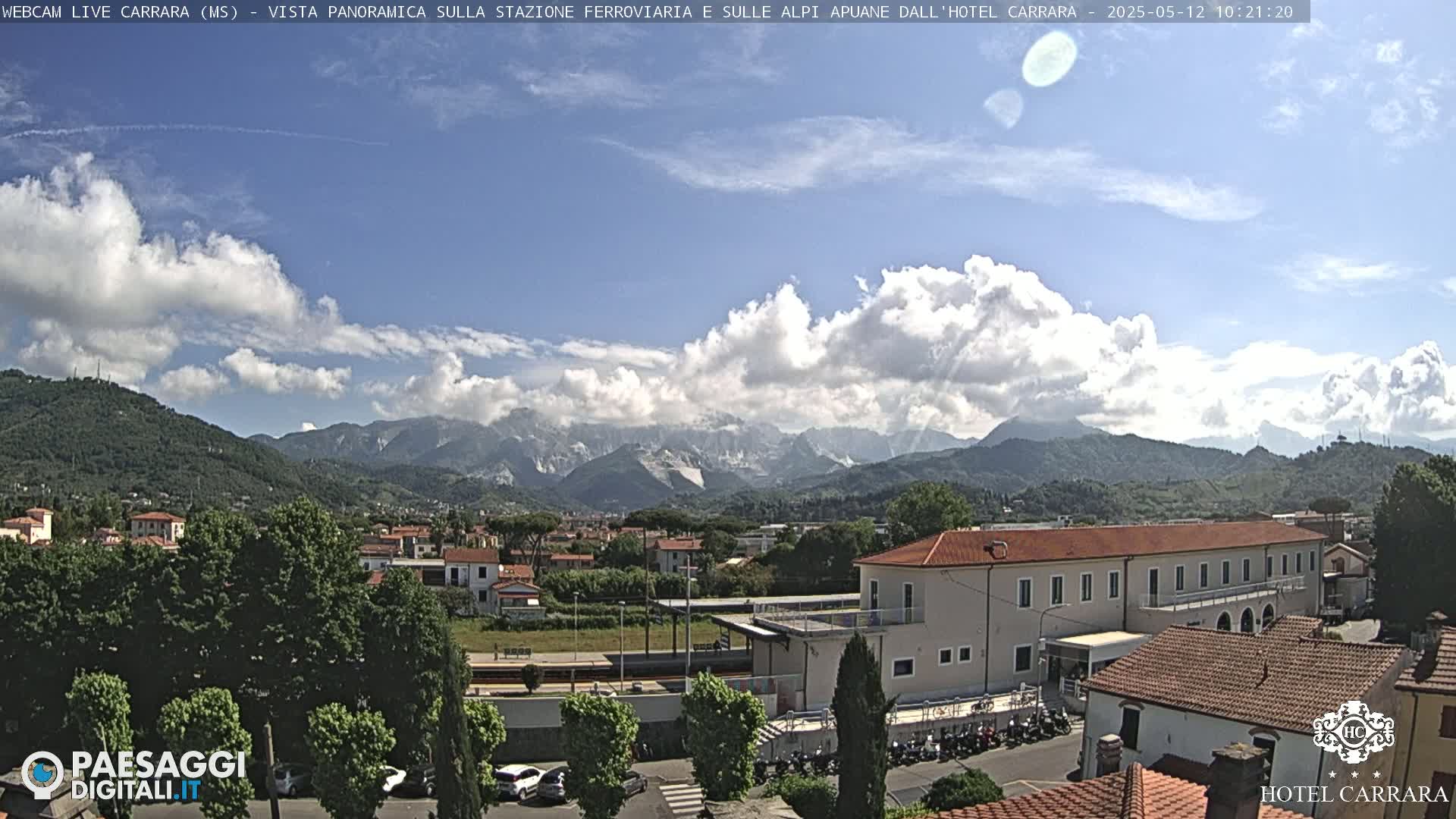 A partly cloudy day reveals a panoramic view of a town nestled at the foot of a mountain range, with a train station and buildings visible in the foreground.