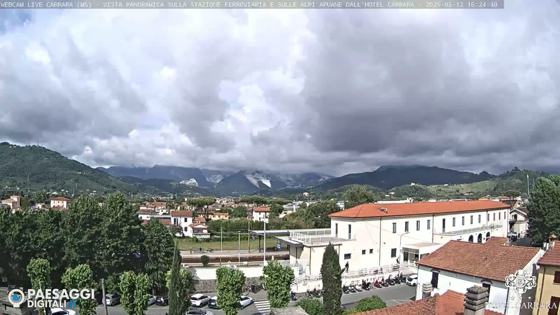 A partly cloudy day reveals a cityscape nestled in a valley between mountains, with a long, low building near the foreground.