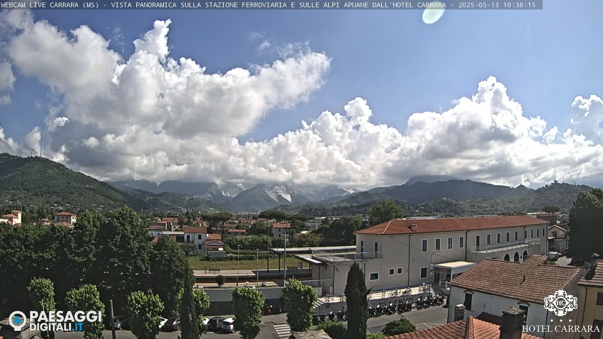 A partly cloudy day reveals a panoramic view of a town nestled in a valley, with mountains in the background and a train station visible in the foreground.