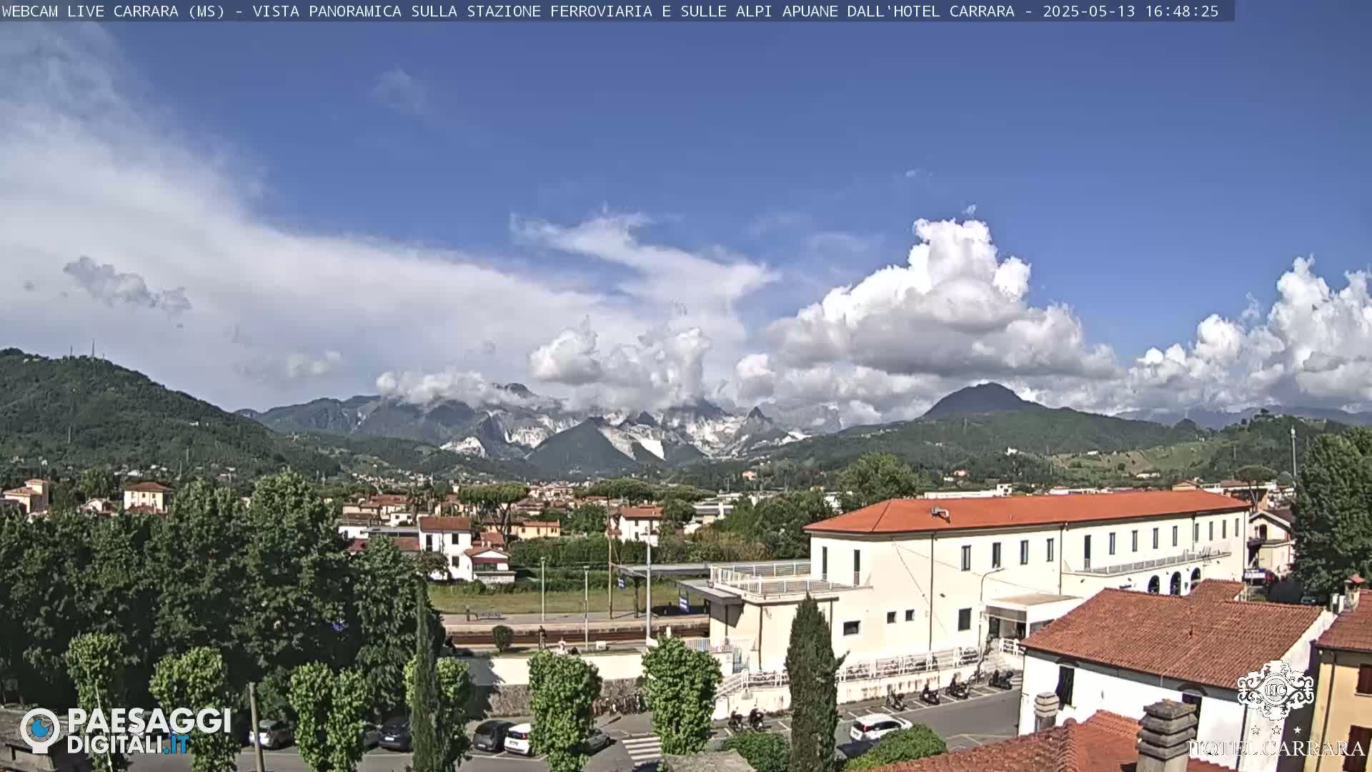 A partly cloudy day reveals a panoramic view of a town nestled in a valley, with mountains in the background and a long, low building in the foreground.