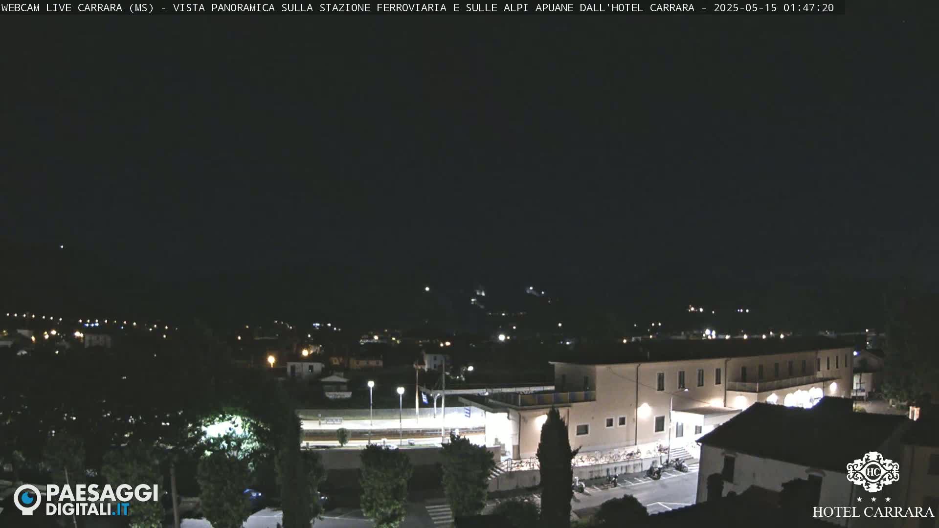 A nighttime, panoramic view of a town, featuring a train station and a large building, possibly a hotel, under a clear, dark sky.