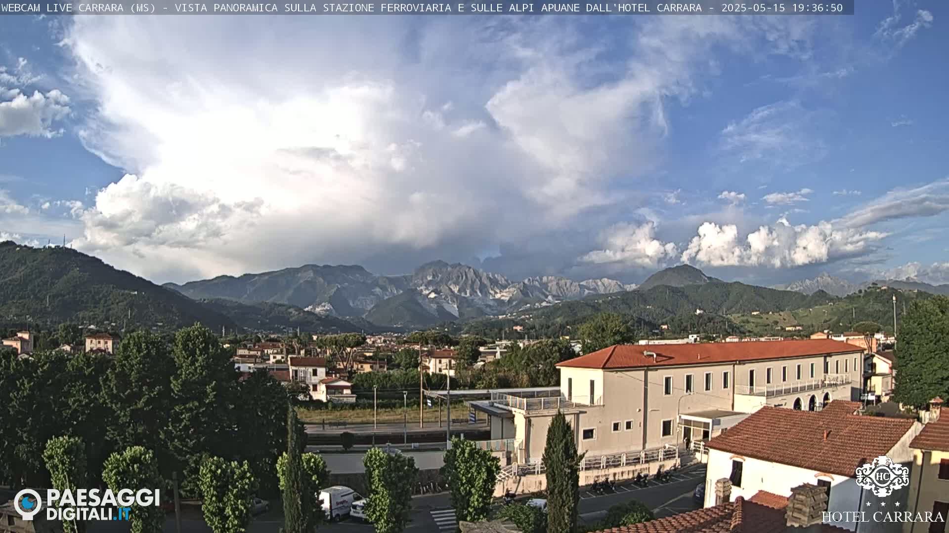 A partly cloudy sky over a town nestled in a valley, with mountains in the background.