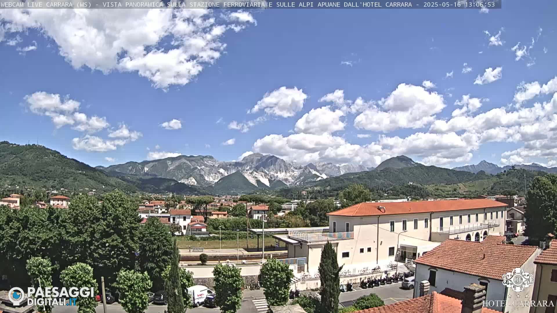 A partly sunny day reveals a view of a town nestled in a valley, with mountains in the background and a long building, possibly a hotel or train station, in the foreground.