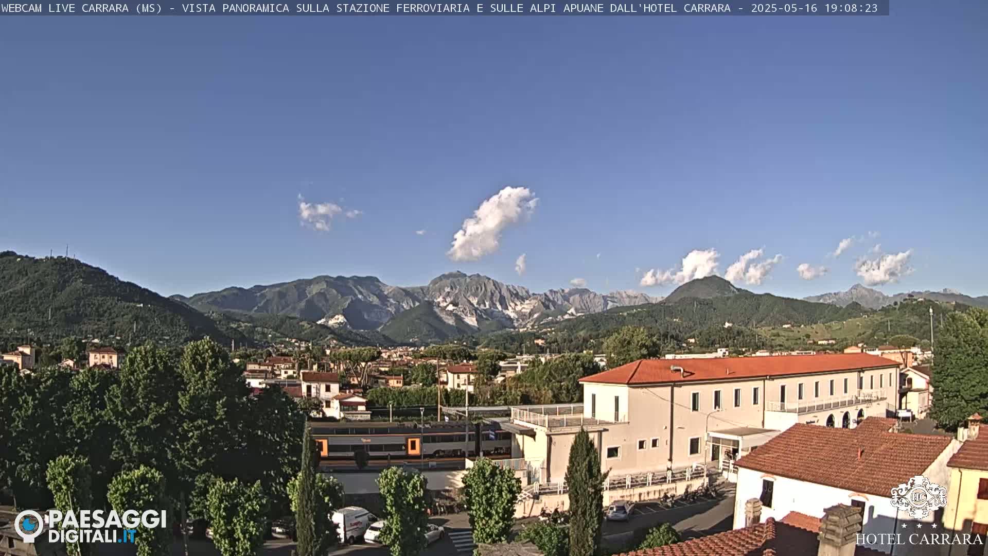 A partly cloudy sunny day reveals a train in front of a long building, with a town and mountains in the background.